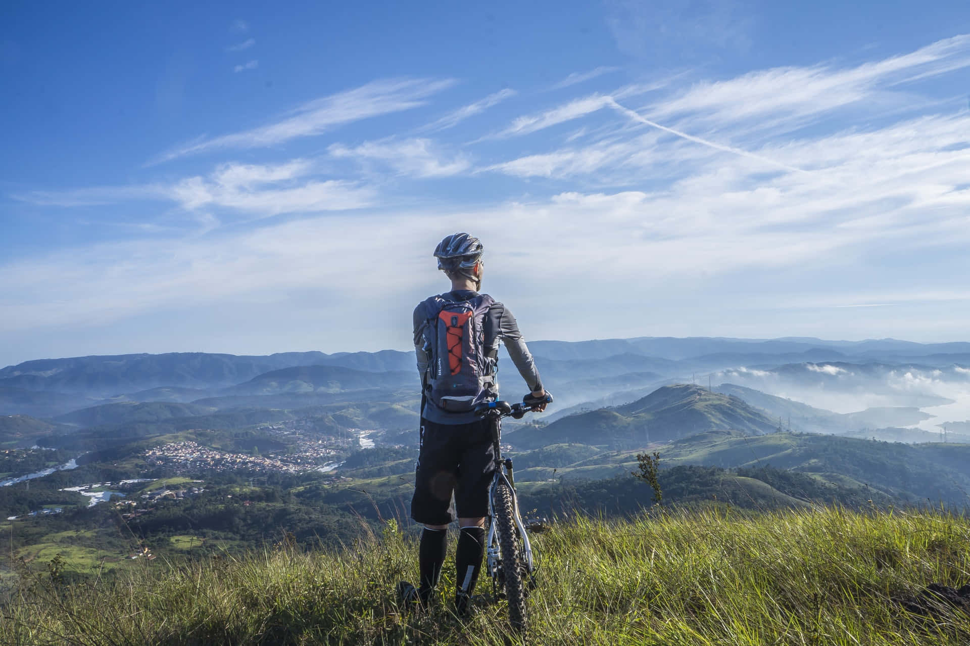 Outdoor Activities 桌布 壁紙 — 一名山地車手站在長滿青草的山脊上，俯瞰著霧氣瀰漫的山谷、起伏的丘陵和遠處的小鎮，天空湛藍明亮。