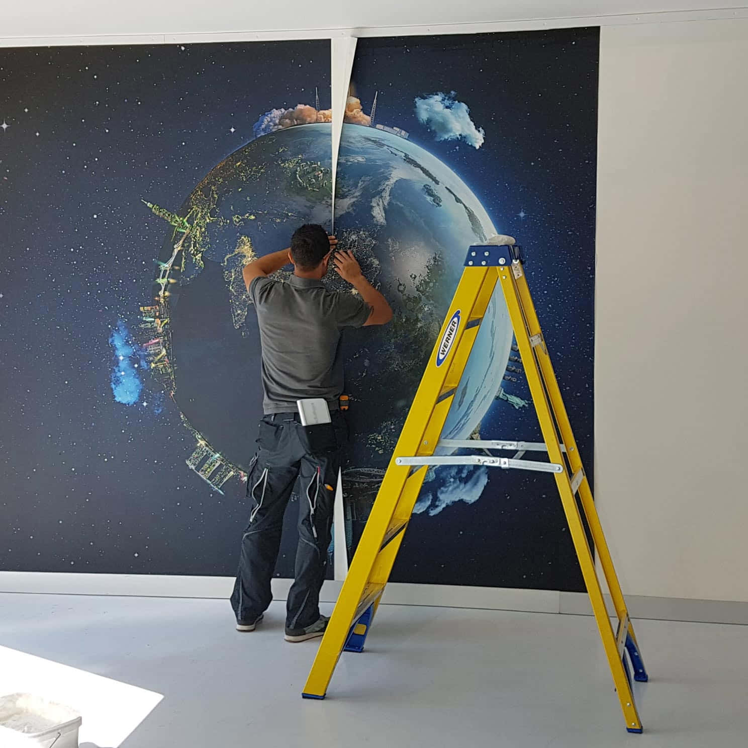 Installation Art 桌布 壁紙 — 一名工人正在安裝一幅大型太空主題牆紙，上面有地球和星星，旁邊有一把黃色梯子，牆壁是素色的。