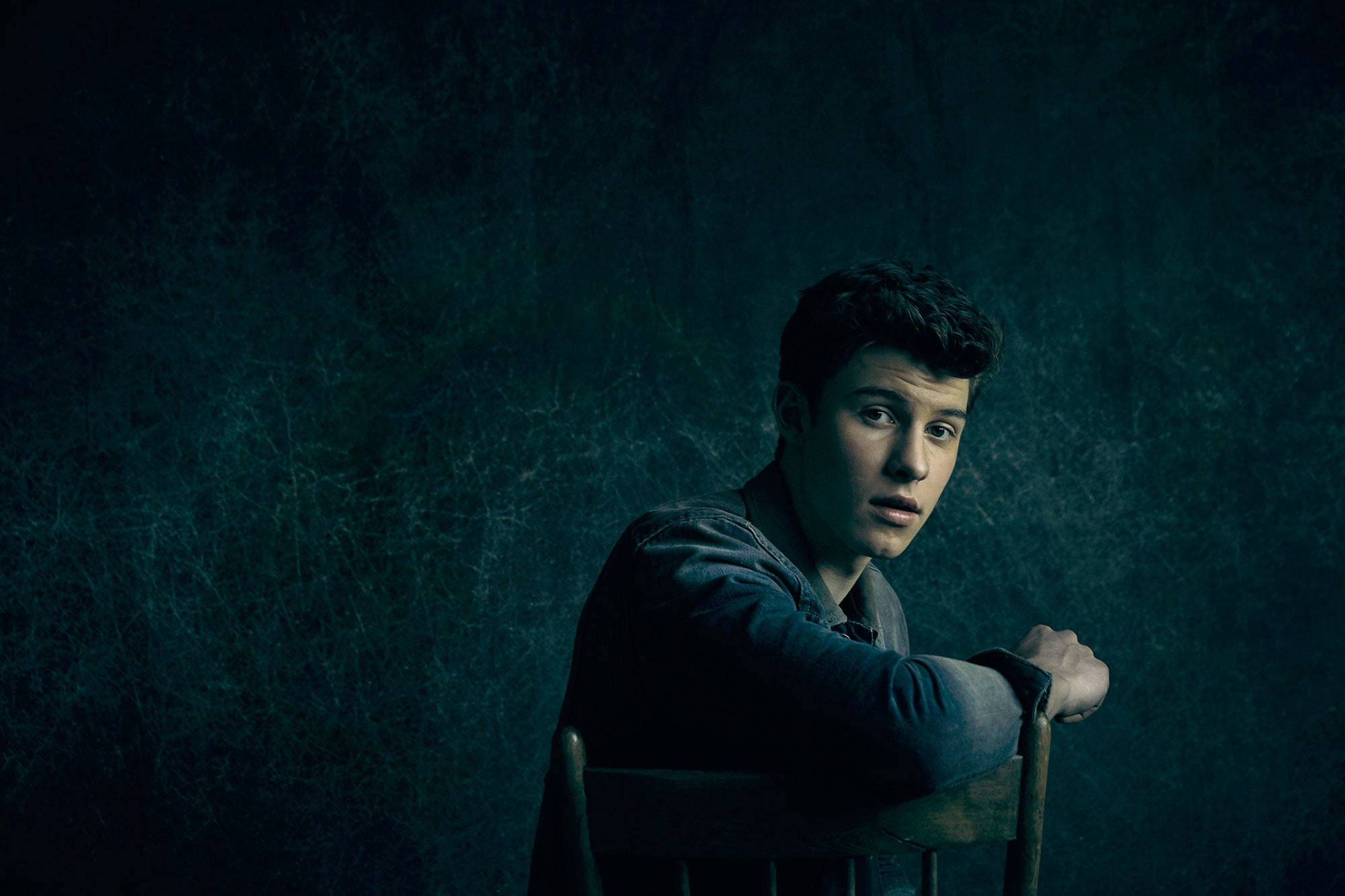 Shawn Mendes《Illuminate》 壁紙 — 一名年輕男子在戲劇性的工作室燈光下，靠在一張木椅上，身體前傾，靠著一面有紋理的深藍綠色牆壁。