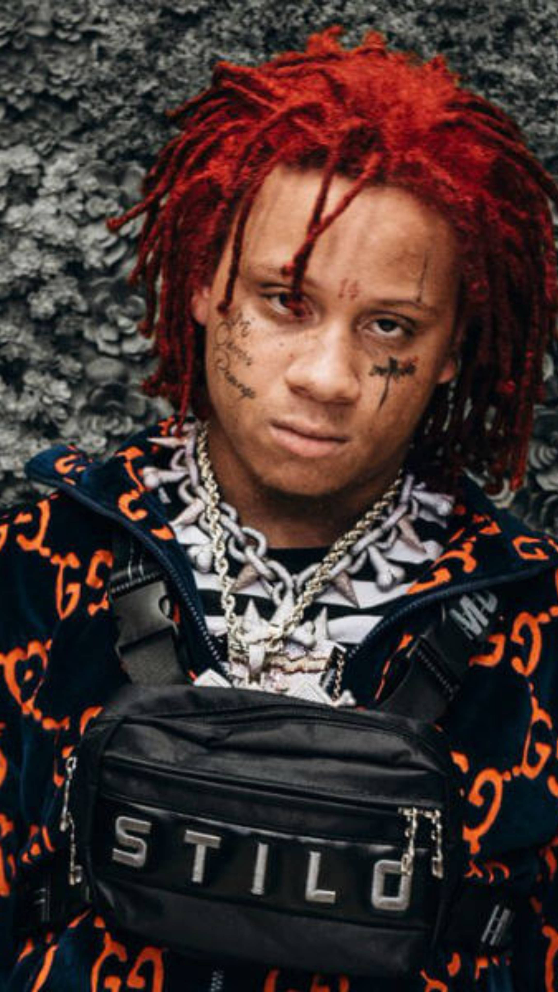 Trippie Redd STILO 壁紙 — 一名年輕男子擁有鮮紅色辮子和臉部紋身，對著紋理灰色牆壁凝視，穿著大膽圖案外套和黑色'STILO'腰包。