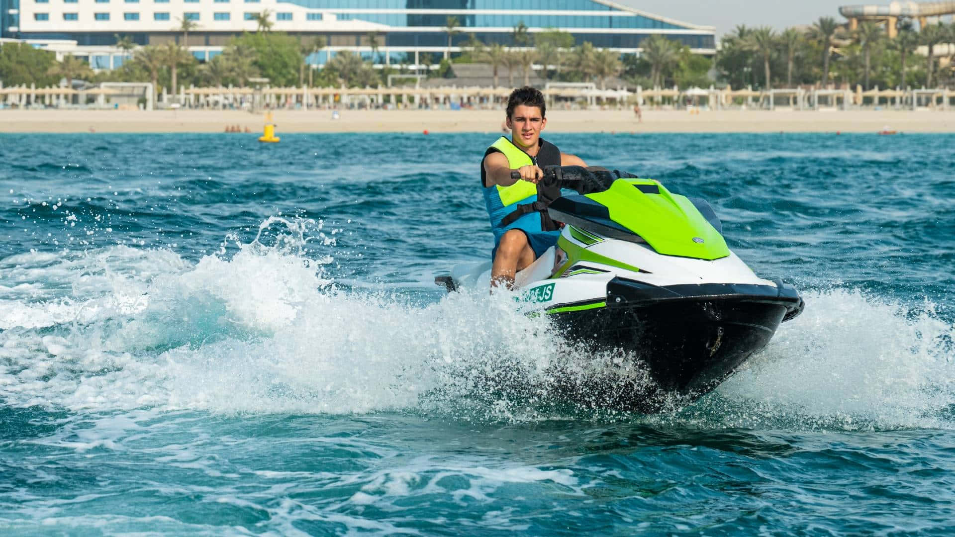 Beach Water Sports 桌布 壁紙 — 一名年輕男子騎著亮綠色水上電單車，在碧綠色的海水上高速行駛，背景是沙灘、棕櫚樹和現代建築。