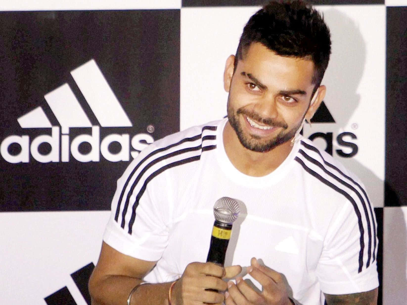 Virat Kohli 桌布 壁紙 — 一名微笑的男子身穿白色adidas襯衫，在品牌背景前手持麥克風。