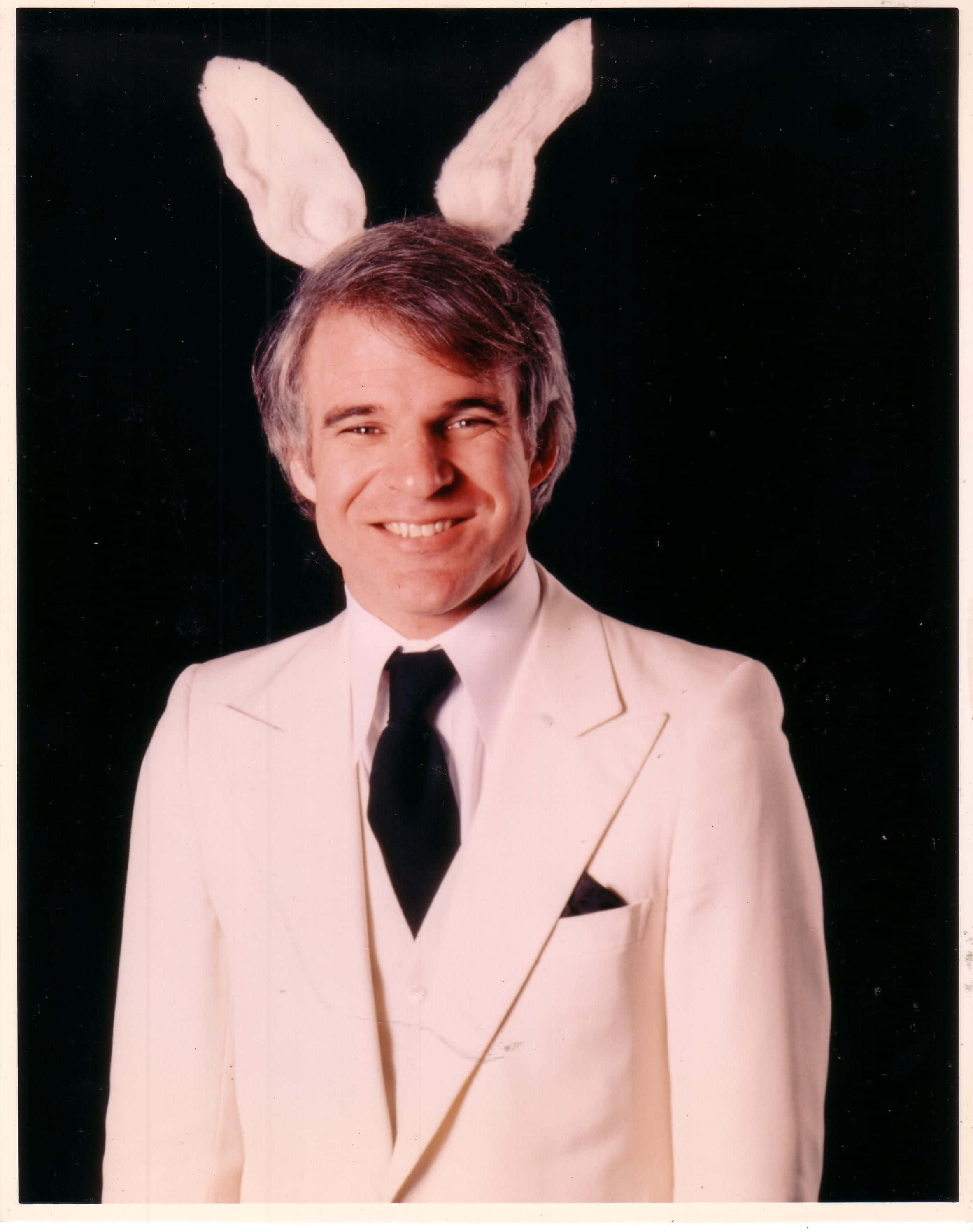 steve martin 桌布 壁紙 — 一名微笑的男子身穿白色西裝和黑色領帶，戴著白色兔耳，背景為黑色。