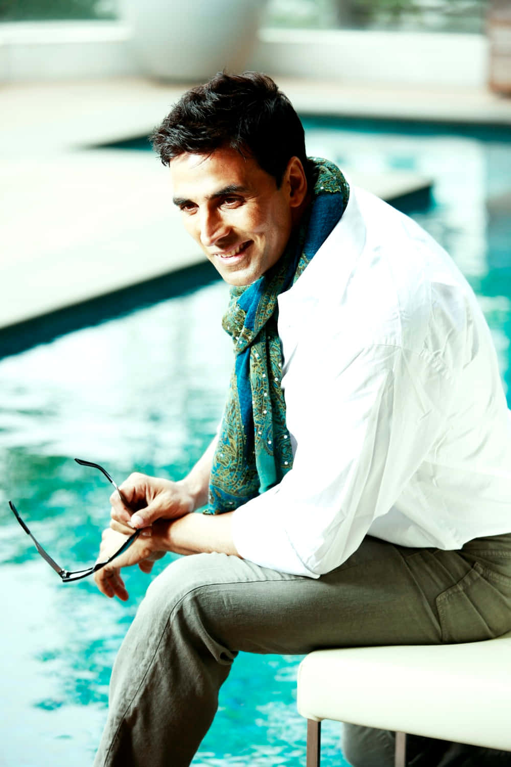 Akshay Kumar 桌布 壁紙 — 一名微笑的男子身穿白襯衫和圖案圍巾，坐在藍綠色泳池旁，手持太陽眼鏡。