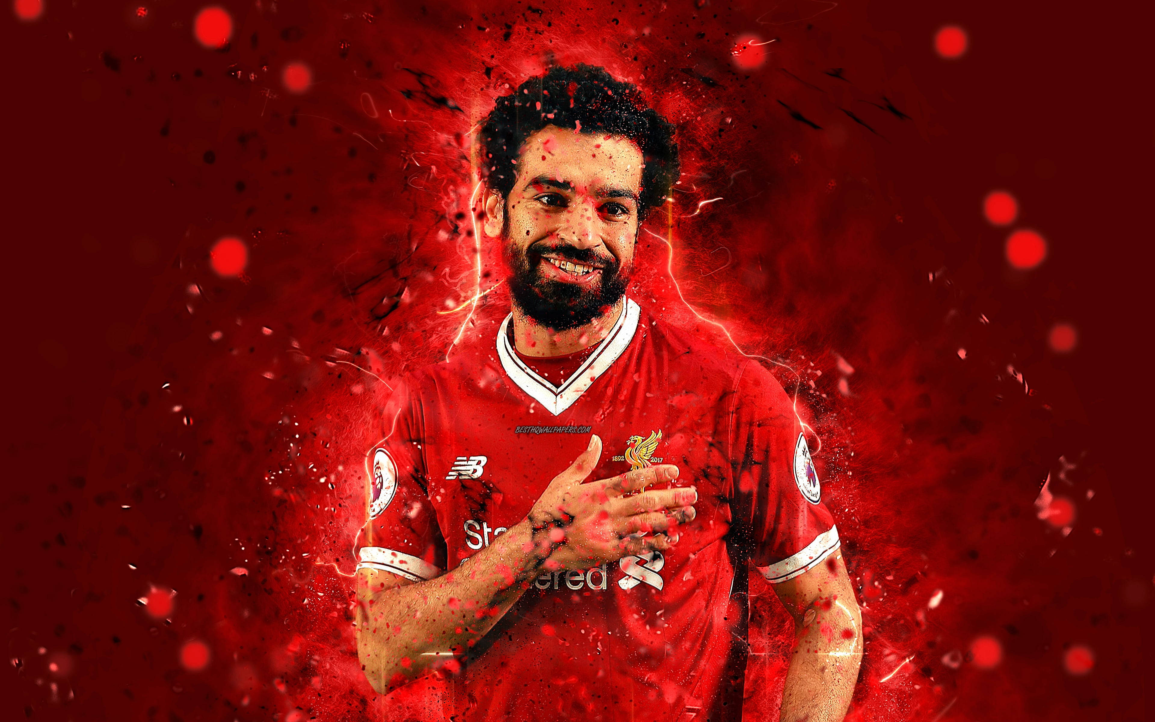 Liverpool Fc 桌布 壁紙 — 一名微笑的足球員身穿紅色利物浦球衣，站在充滿電火花和浮游粒子的動態紅色背景前。
