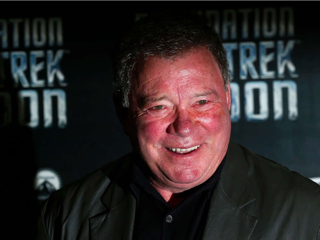 william shatner 桌布 壁紙 — 一名微笑的黑外套男子站在帶有『Star Trek』字樣的深色背景前。