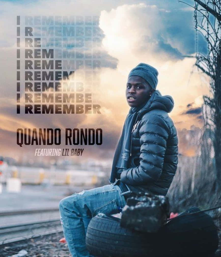 Quando Rondo 桌布 壁紙 — 一名戴冷帽穿羽絨外套的年輕男子坐在火車軌道旁的輪胎上，背景是戲劇性的日落天空，並有重複的文字疊加。