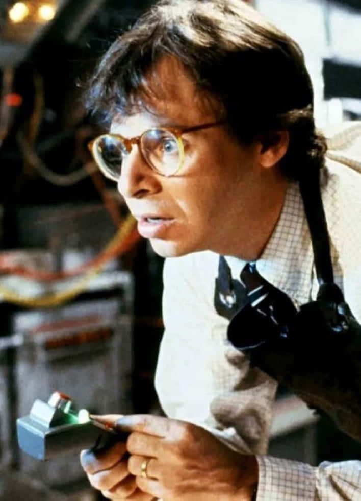 rick moranis 桌布 壁紙 — 一名戴圓框眼鏡、身穿格仔恤衫嘅男人喺一個佈滿電線同設備嘅凌亂實驗室入面揸住一個發光裝置。