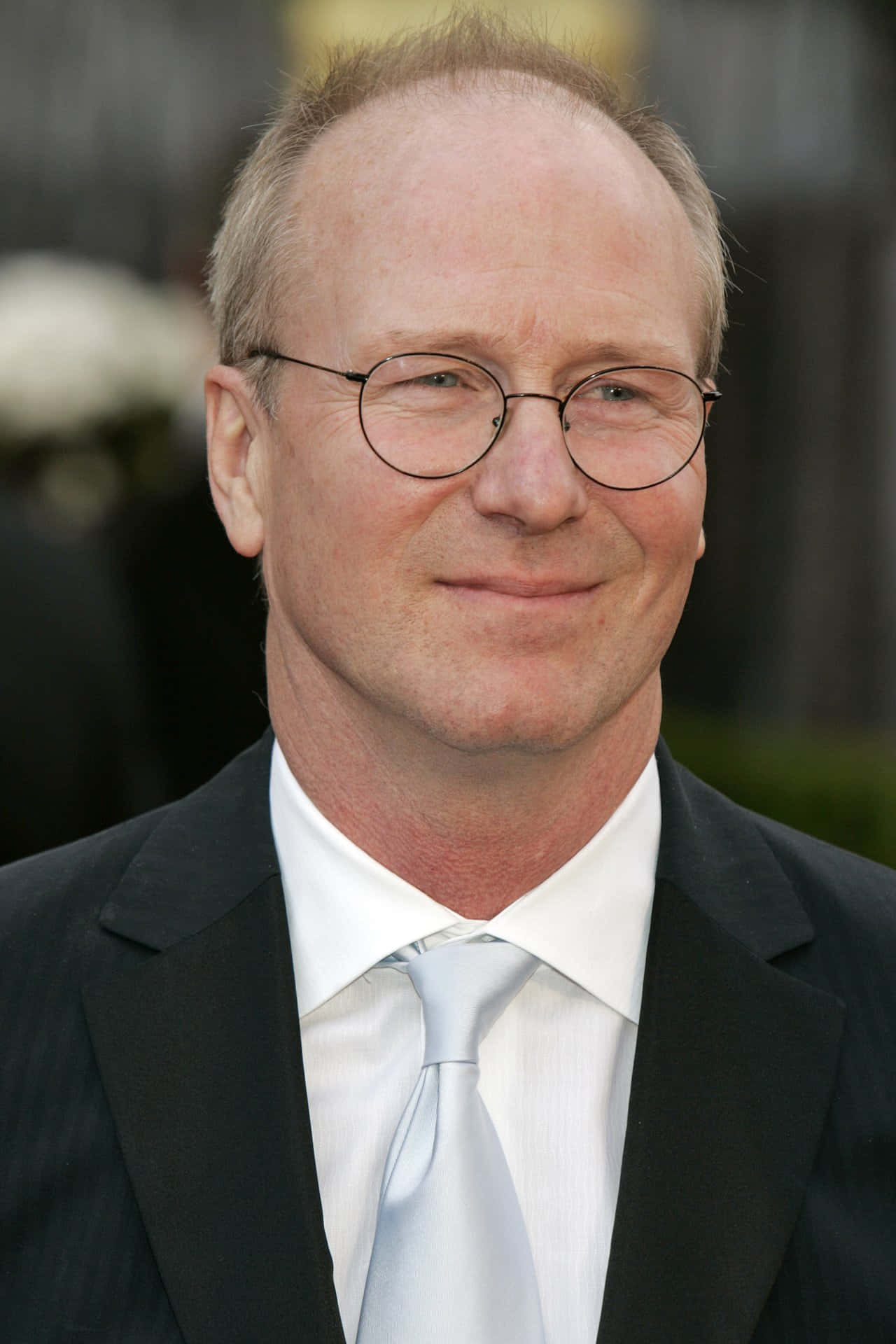 William Hurt 桌布 壁紙 — 一名戴圓框眼鏡、身穿黑色西裝及淺藍色領帶的男士特寫肖像，背景模糊。
