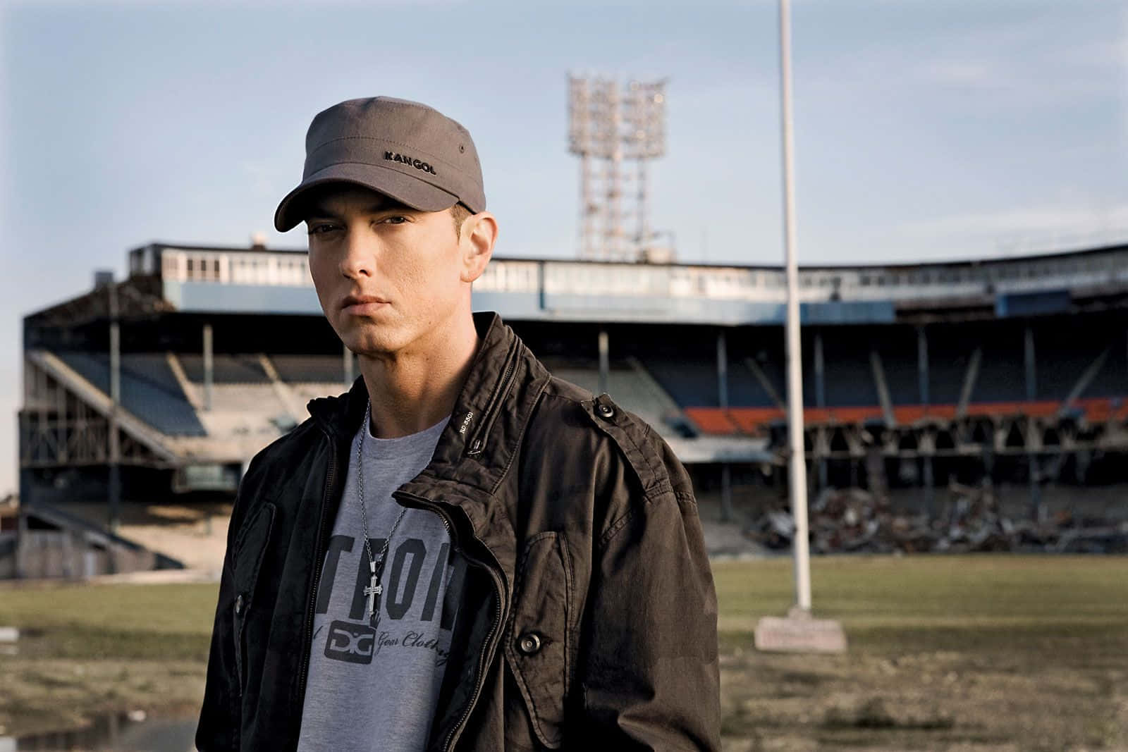 Eminem 桌布 壁紙 — 一名戴帽子和穿夾克的男子在晴朗的天空下莊嚴地站在空蕩蕩的體育場前。