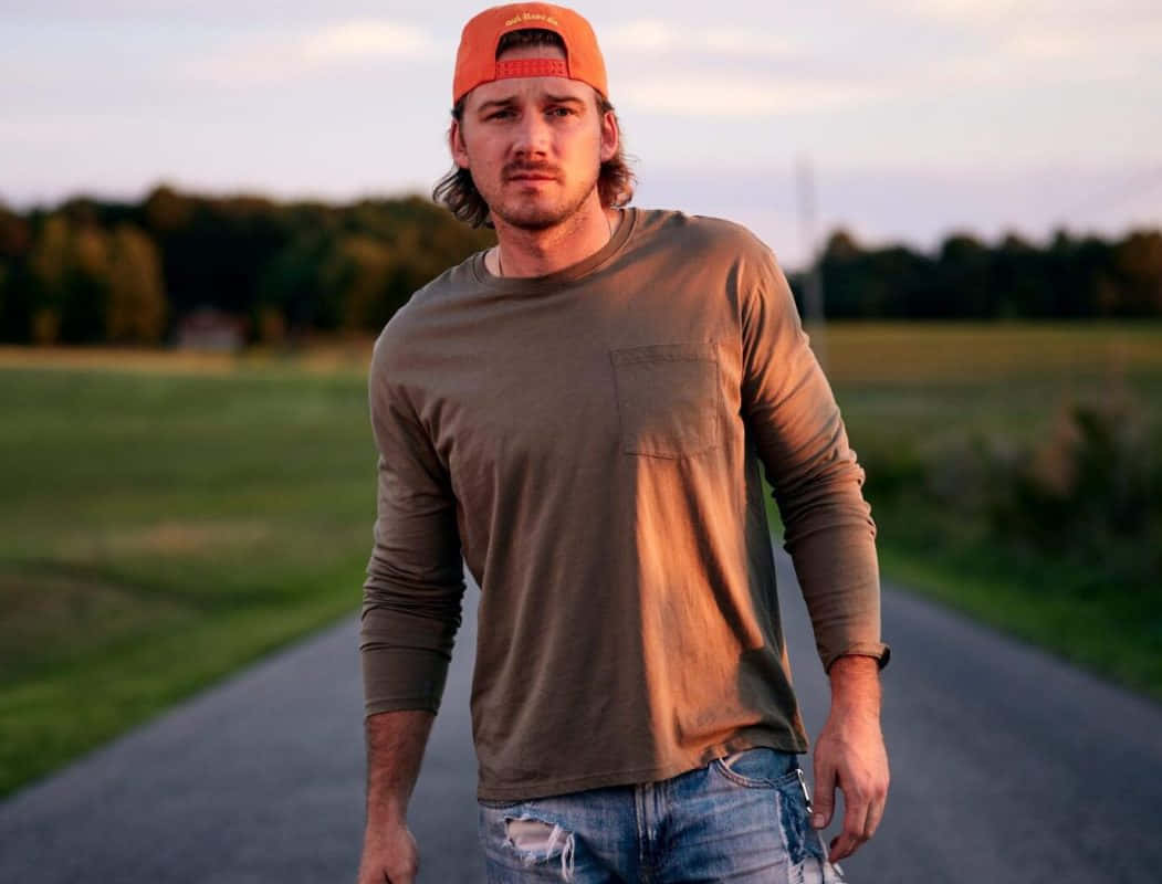 Morgan Wallen 桌布 壁紙 — 一名戴橙色帽、穿棕色衫的男子在黃金時刻沿著鄉村道路行走，身後有樹木和田野。