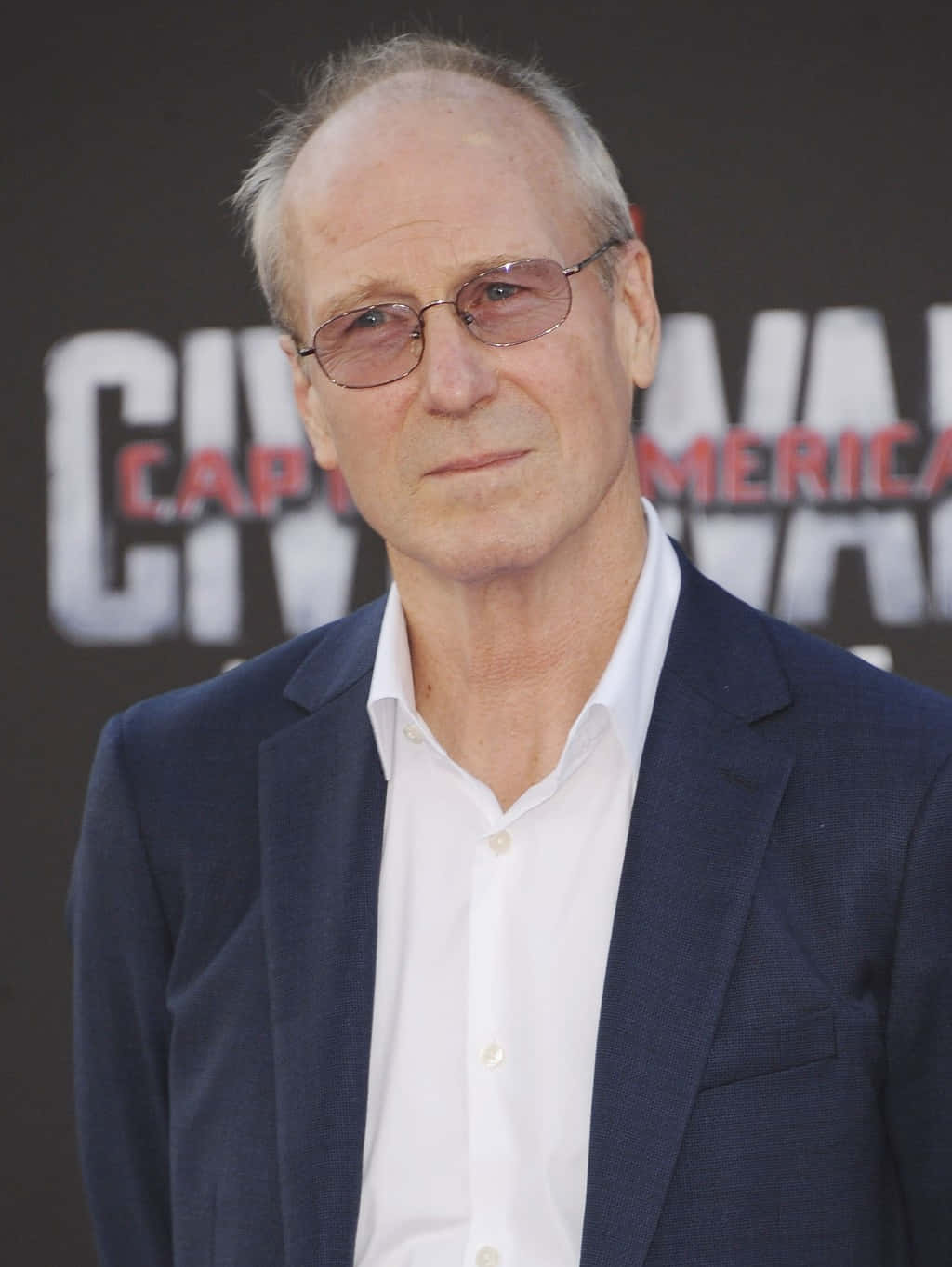 William Hurt 桌布 壁紙 — 一名戴眼鏡、身穿海軍藍西裝外套的男子站在紅地毯活動的《美國隊長》電影背景前。