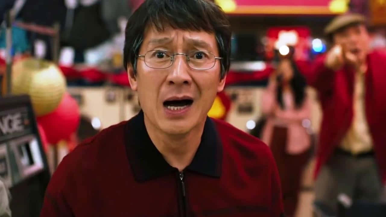 Ke Huy Quan 桌布 壁紙 — 一名戴眼鏡、身穿紅色外套的男子在熱鬧的節日室內環境中張開嘴巴，震驚地凝視。