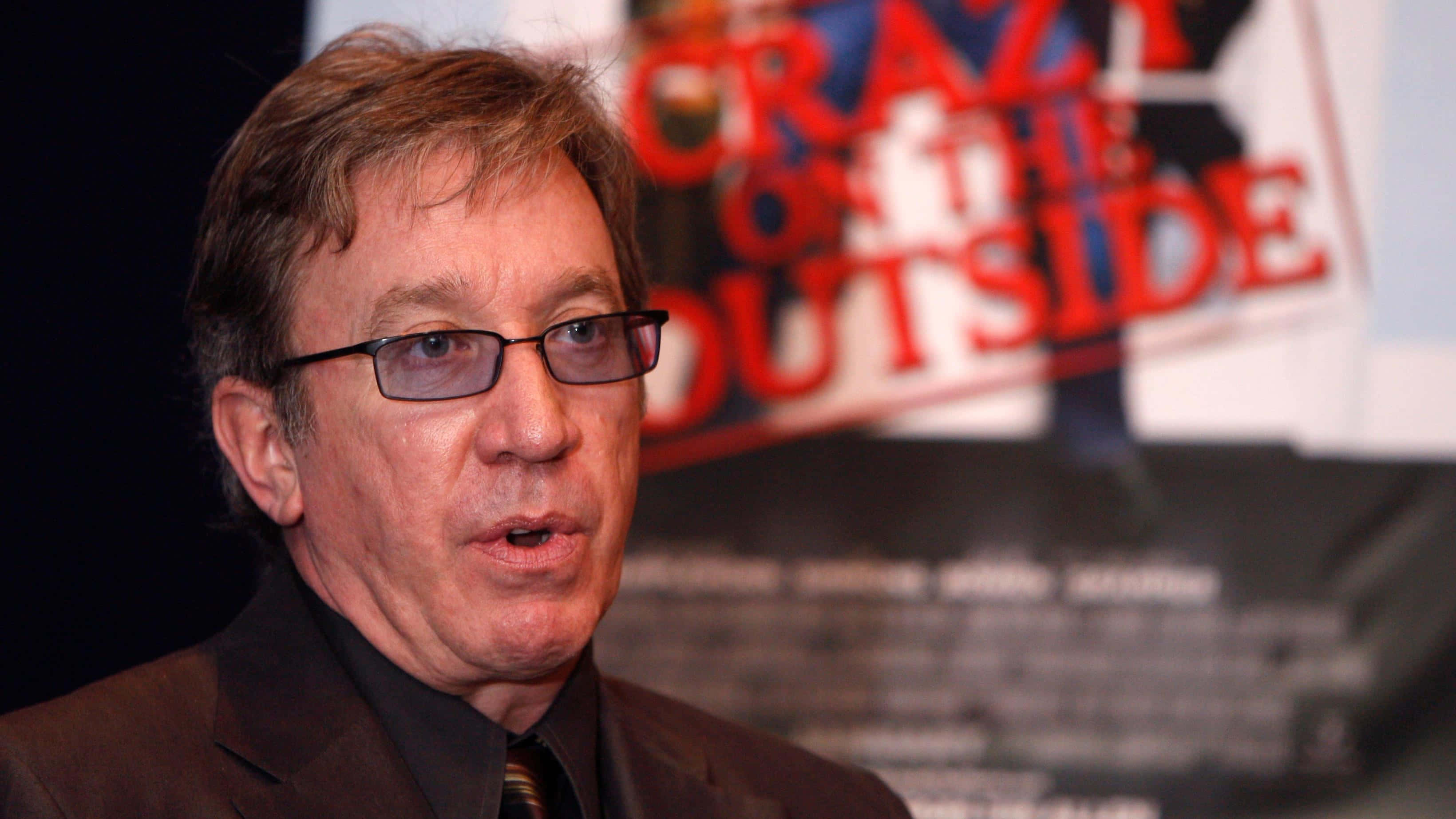 Tim Allen 桌布 壁紙 — 一名戴眼鏡的男子在電影《Crazy on the Outside》海報後方的新聞發布會上發言。