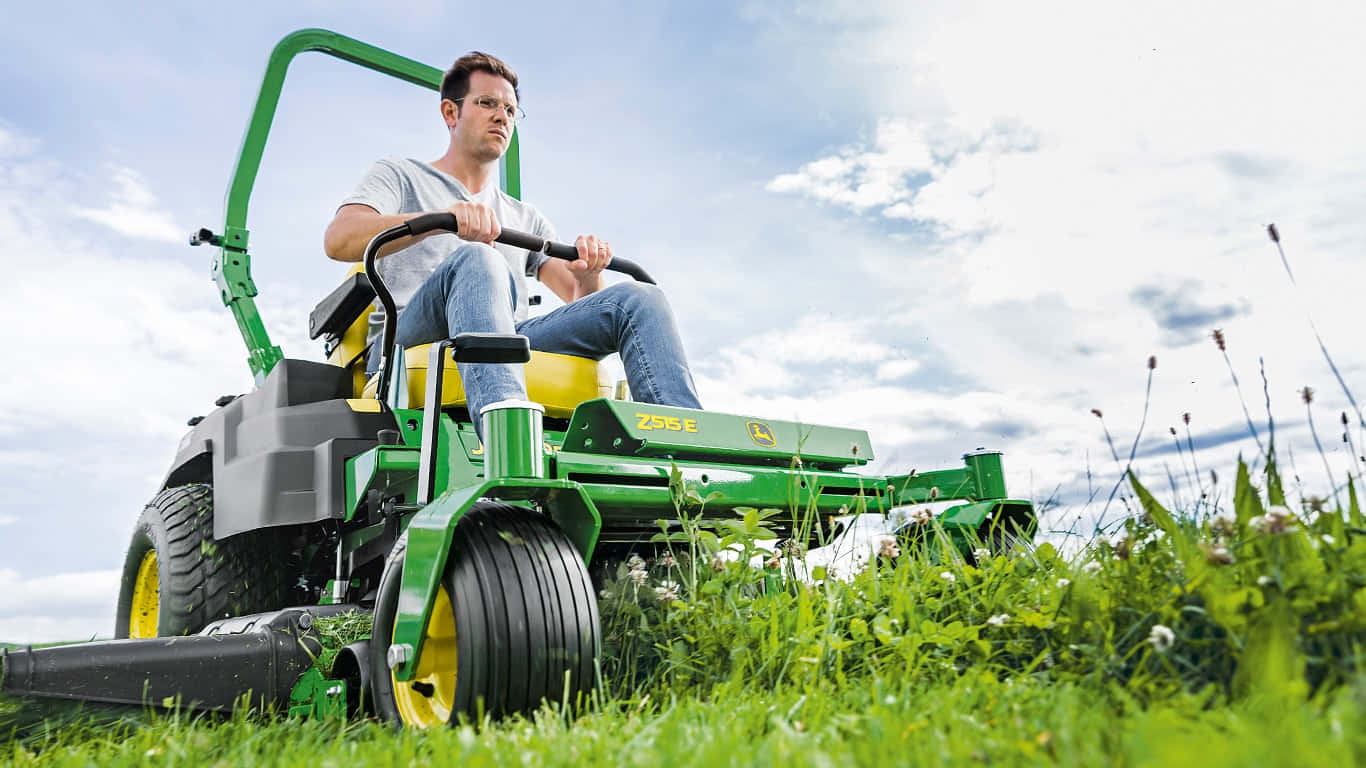 John Deere 桌布 壁紙 — 一名戴眼鏡的男子駕駛綠色John Deere零轉向割草機，在陽光普照的草地上行駛，天空部分多雲。