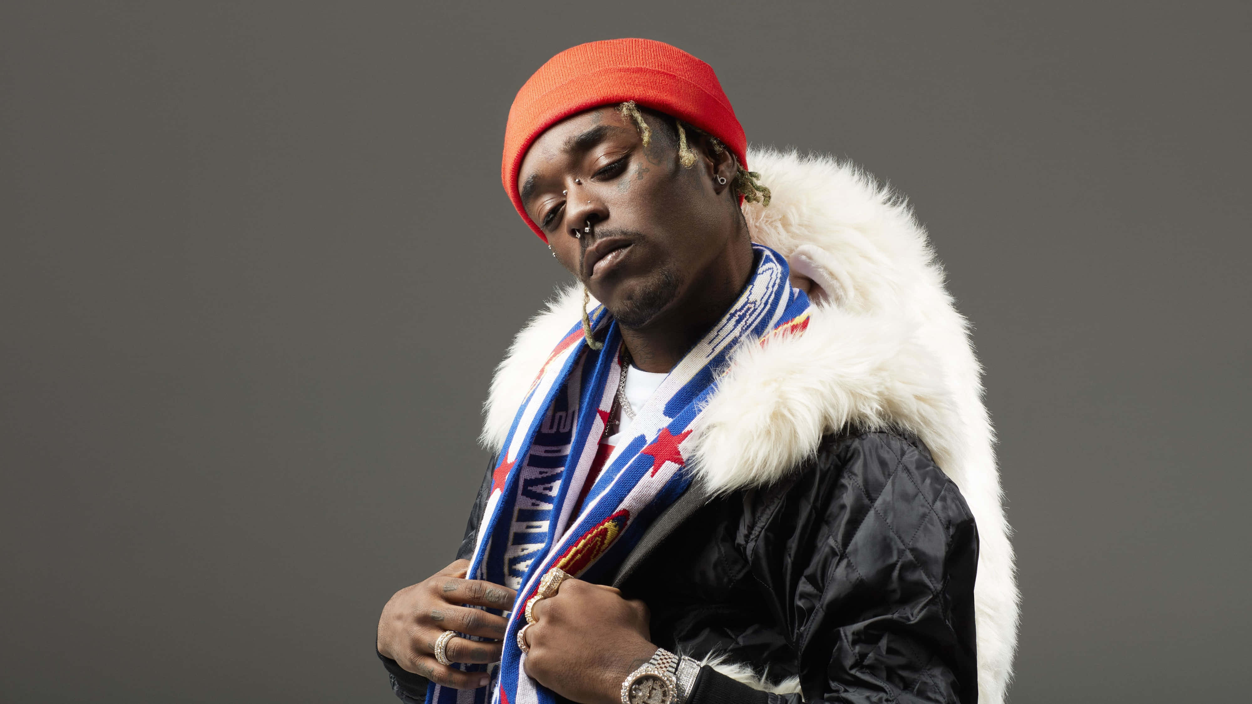 Lil Uzi Vert 桌布 壁紙 — 一名戴紅色冷帽、穿毛領外套的男子，在純灰色背景前閉眼擺姿勢。