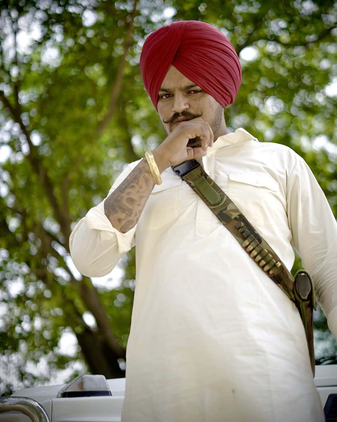 Sidhu Moose Wala 桌布 壁紙 — 一名戴紅色頭巾、身穿白衫的男子，肩上掛著步槍帶，手靠近下巴，站在戶外，背景是綠樹。