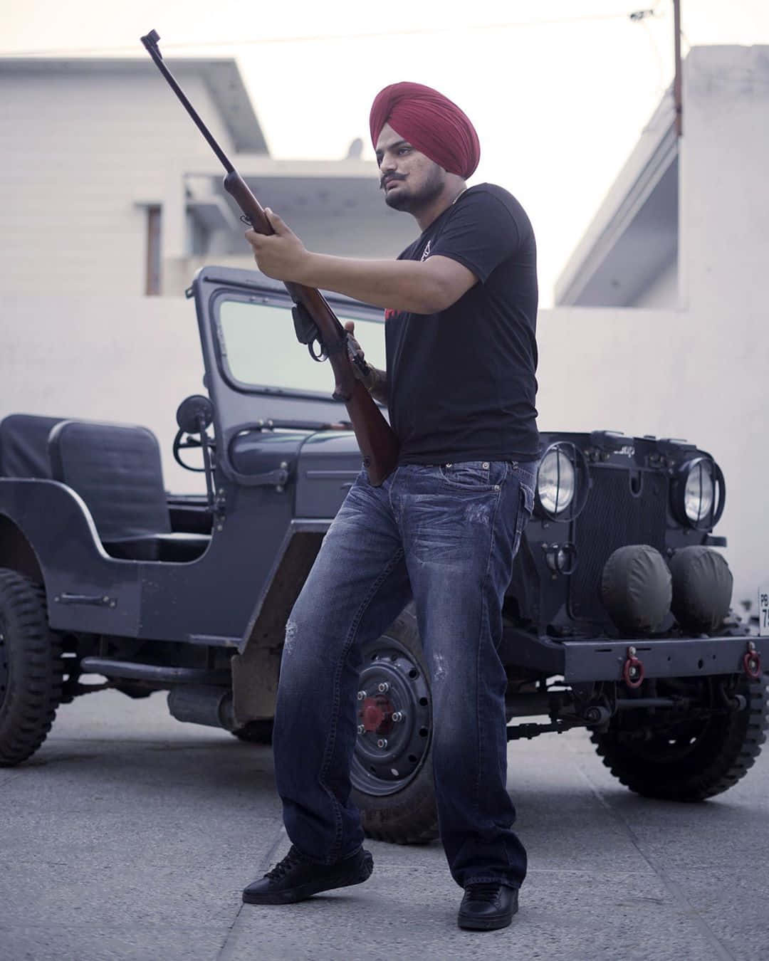 Sidhu Moose Wala 桌布 壁紙 — 一名戴紅色頭巾的男子在陽光明媚的城市庭院中，站在一輛堅固的軍用吉普車旁，手持步槍。