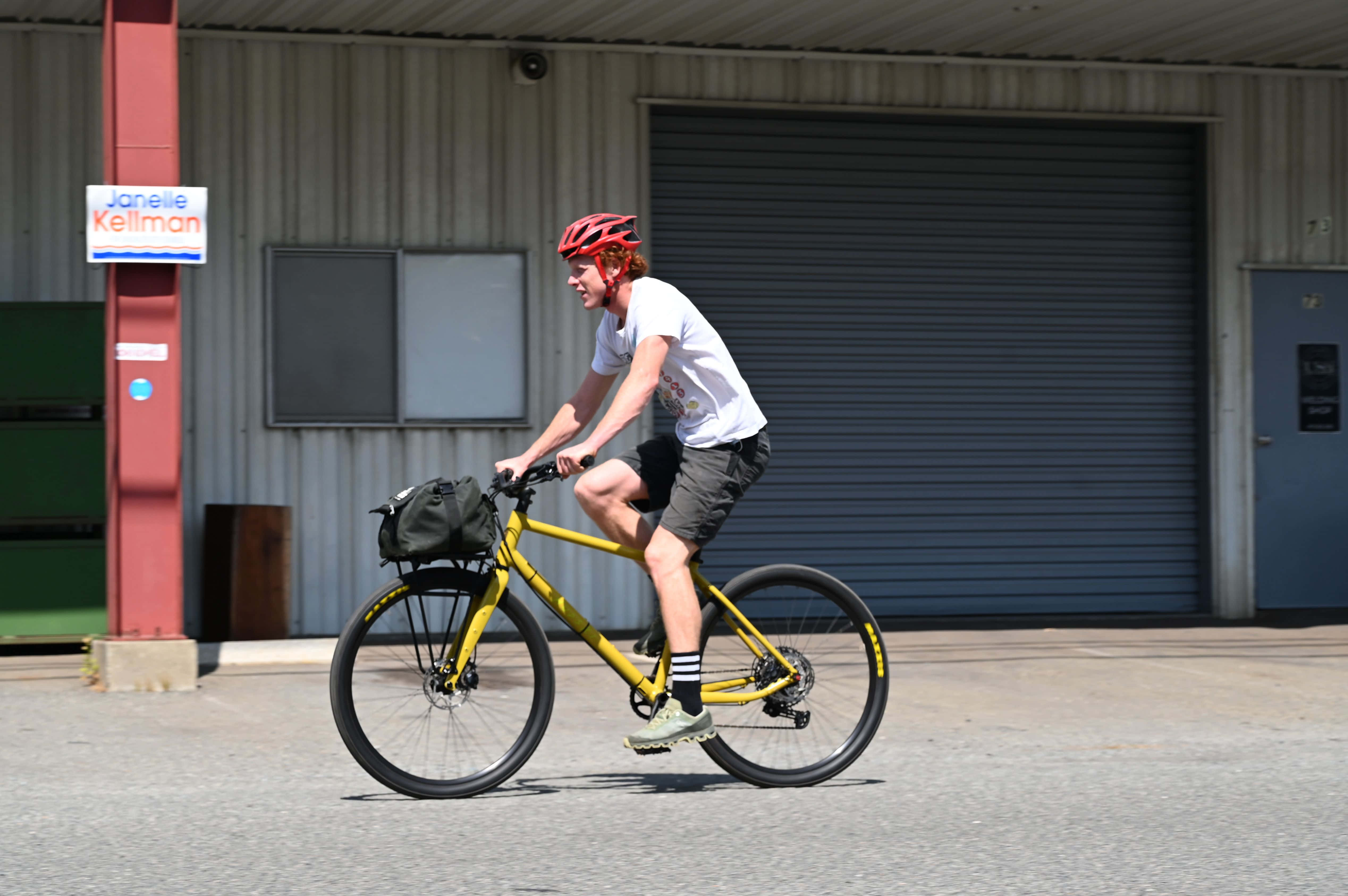 Bicycle 桌布 壁紙 — 一名戴紅色頭盔的騎單車者在晴朗日光下，踩著鮮黃色單車，經過一座波紋金屬建築物前的鋪砌空地。