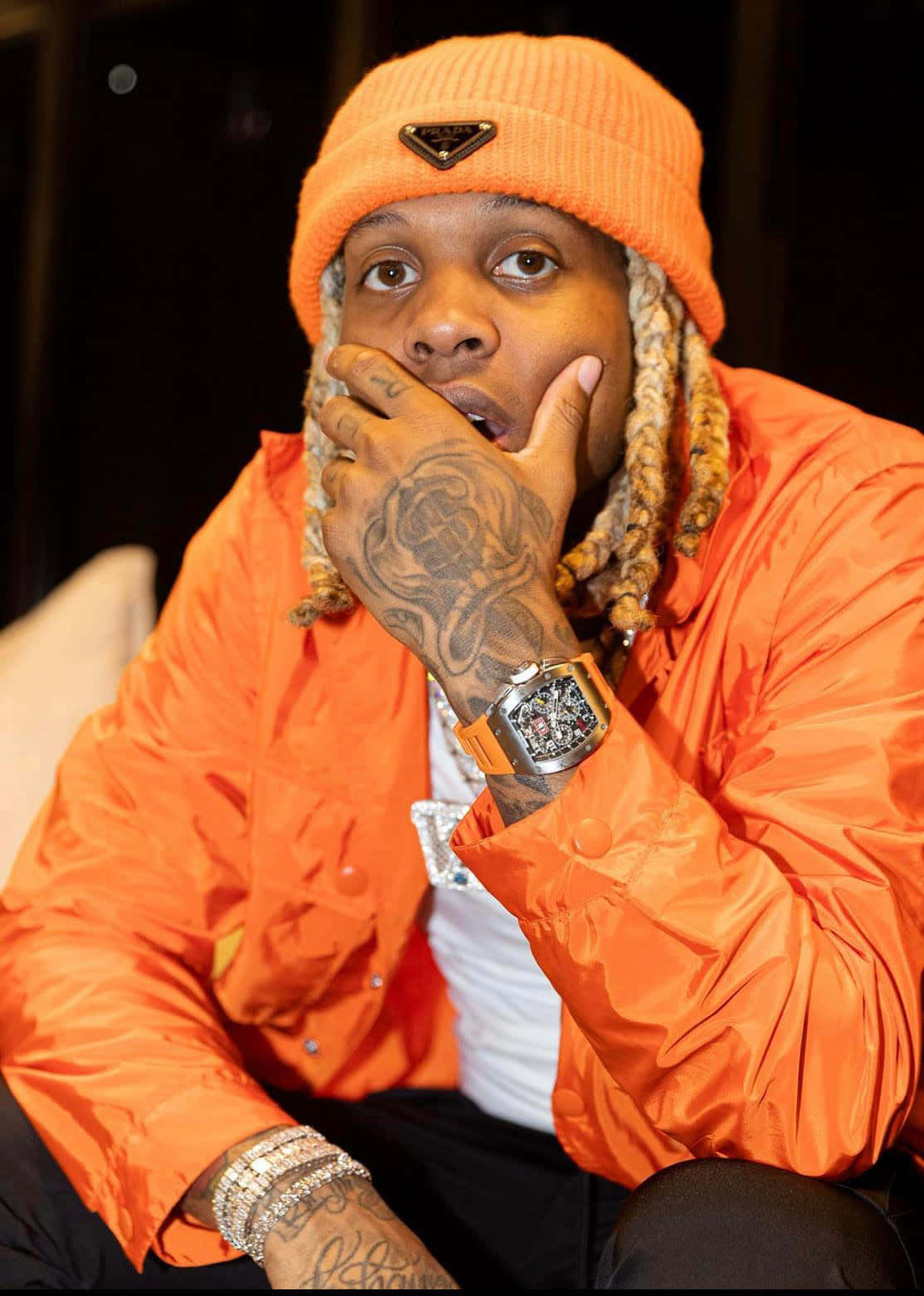 lil durk 桌布 壁紙 — 一名戴著亮橙色冷帽和相配外套的男子，將有紋身的手放在臉旁，佩戴奢華珠寶擺姿勢。
