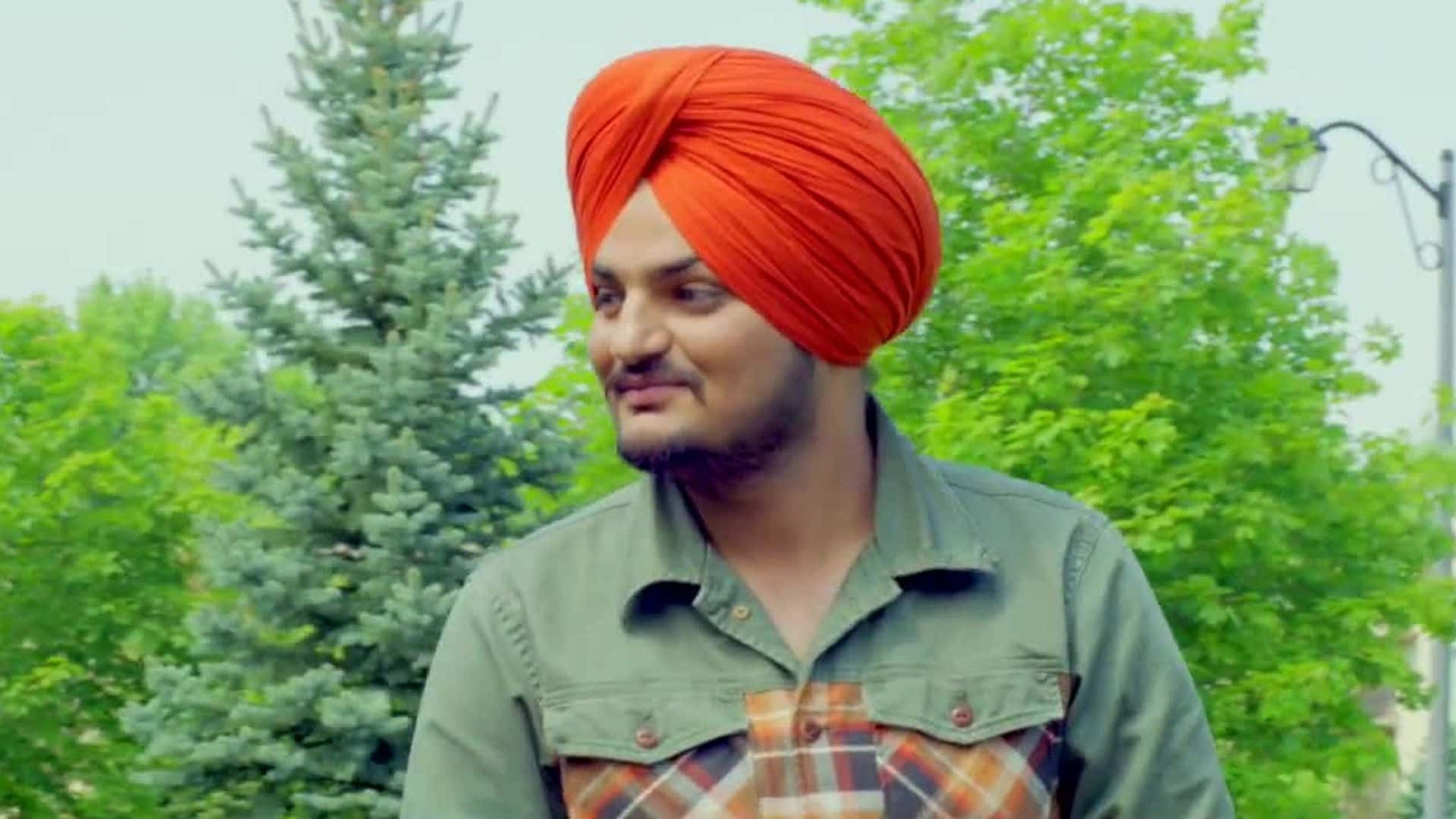 Sidhu Moose Wala 桌布 壁紙 — 一名戴著亮橙色頭巾的男子在戶外，在茂密的綠樹和復古路燈中溫柔地微笑。