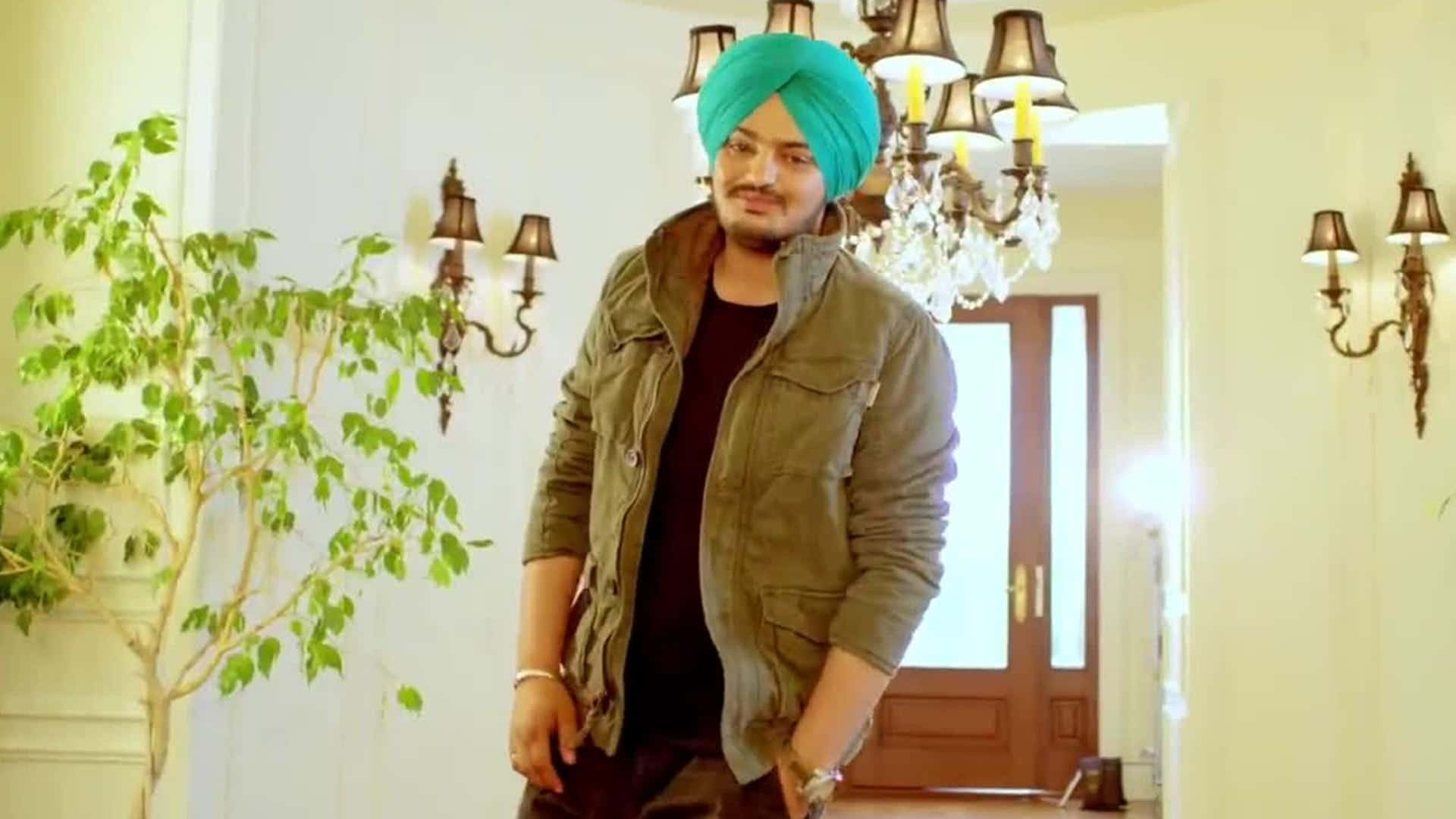 Sidhu Moose Wala 桌布 壁紙 — 一名戴著亮青色頭巾的男子自信地站在一個溫暖照明的走廊中，走廊裝飾著水晶吊燈和綠色植物。