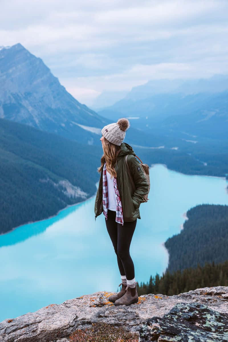 Winter Hiking 桌布 壁紙 — 一名戴著冷帽和外套的女子站在岩石懸崖上，俯瞰著迷霧山巒之間令人驚嘆的碧綠湖泊。