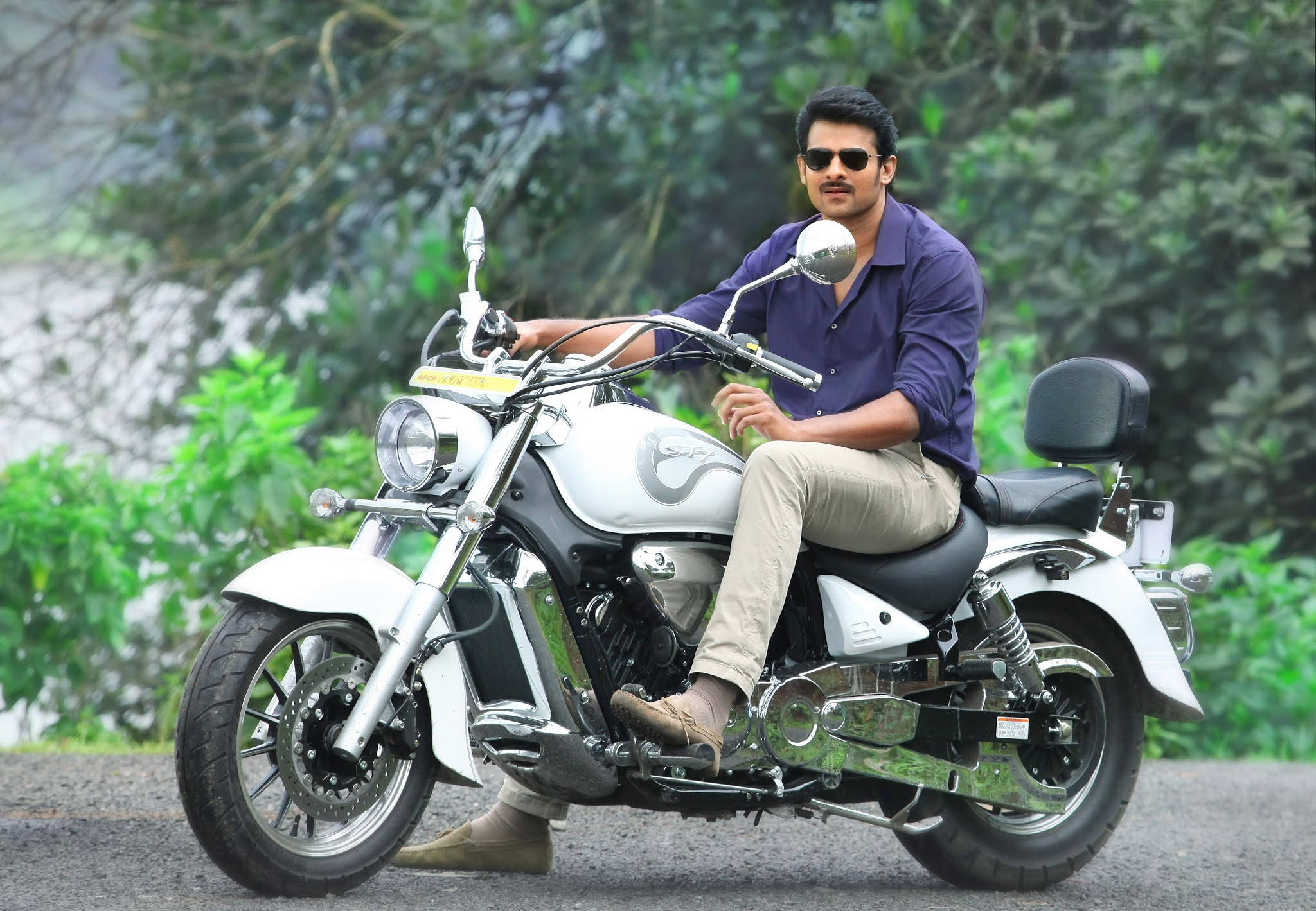Prabhas Mirchi 桌布 壁紙 — 一名戴著太陽眼鏡、身穿紫色襯衫的男子自信地坐在一輛停在碎石路上的白色巡航電單車上，周圍是茂密的綠色植物。