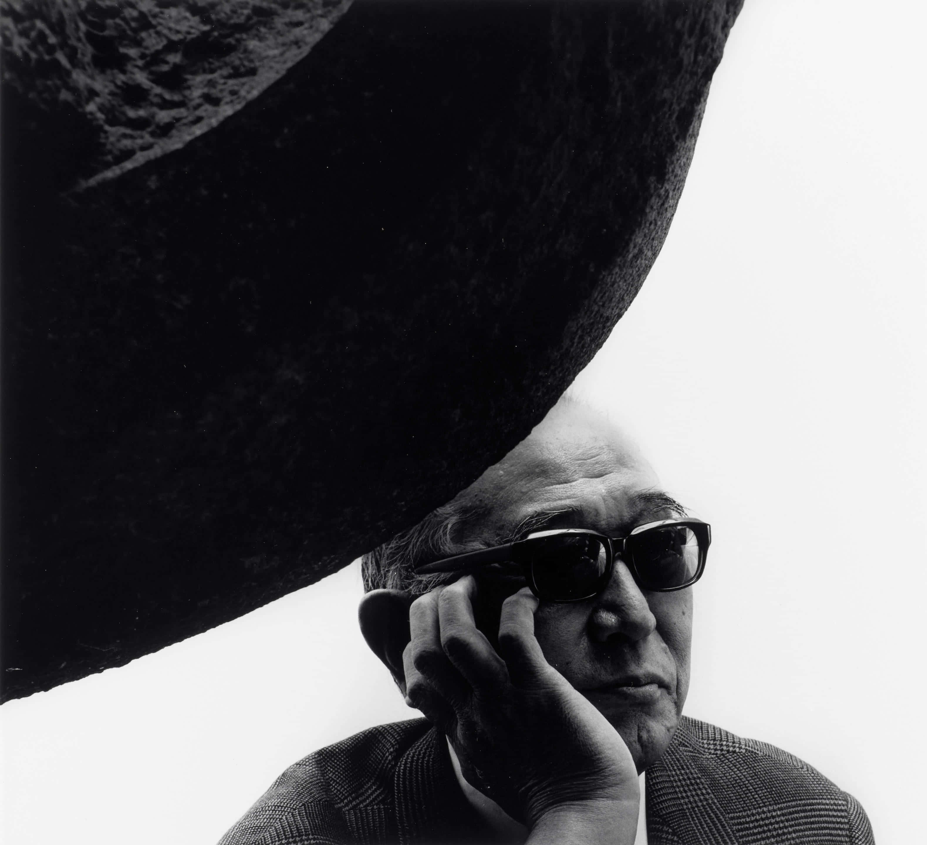 Akira Kurosawa 桌布 壁紙 — 一名戴著太陽眼鏡的男子，在純白背景前，將一塊巨大的深色岩石高舉過頭，形成一張戲劇性的黑白肖像照。