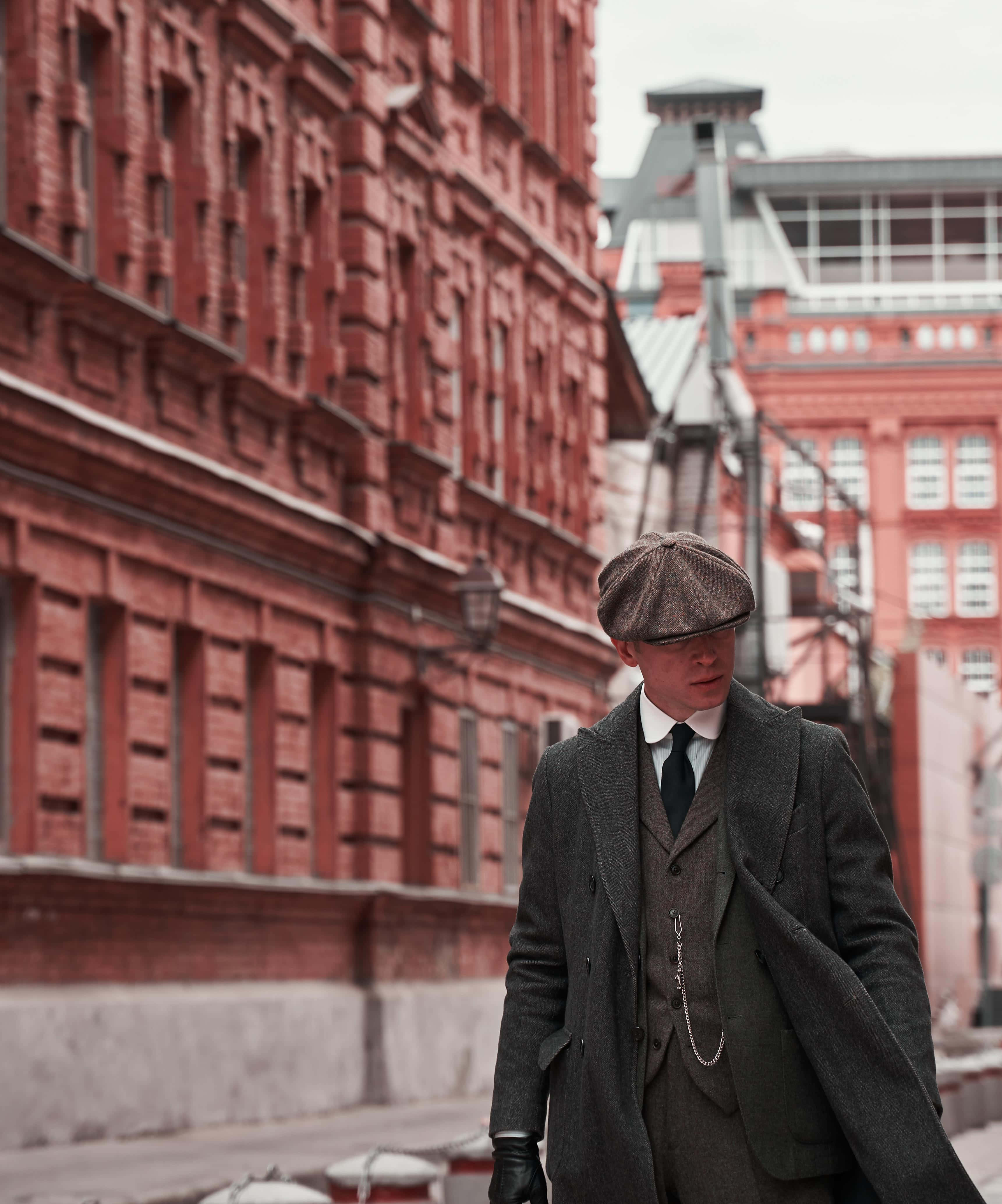 Peaky Blinders 桌布 壁紙 — 一名戴著復古平頂帽、穿著大衣的男子，在陰天下一條兩旁是紅磚建築的狹窄街道上行走。