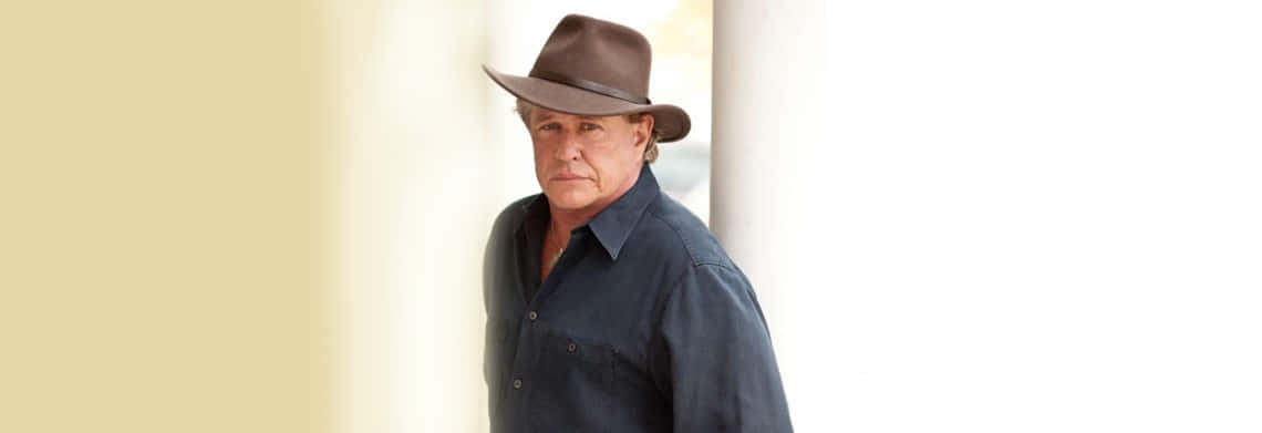 Tom Berenger 桌布 壁紙 — 一名戴著棕色軟呢帽、身穿深色襯衫的男子，站在明亮的柱狀背景前，光線柔和。