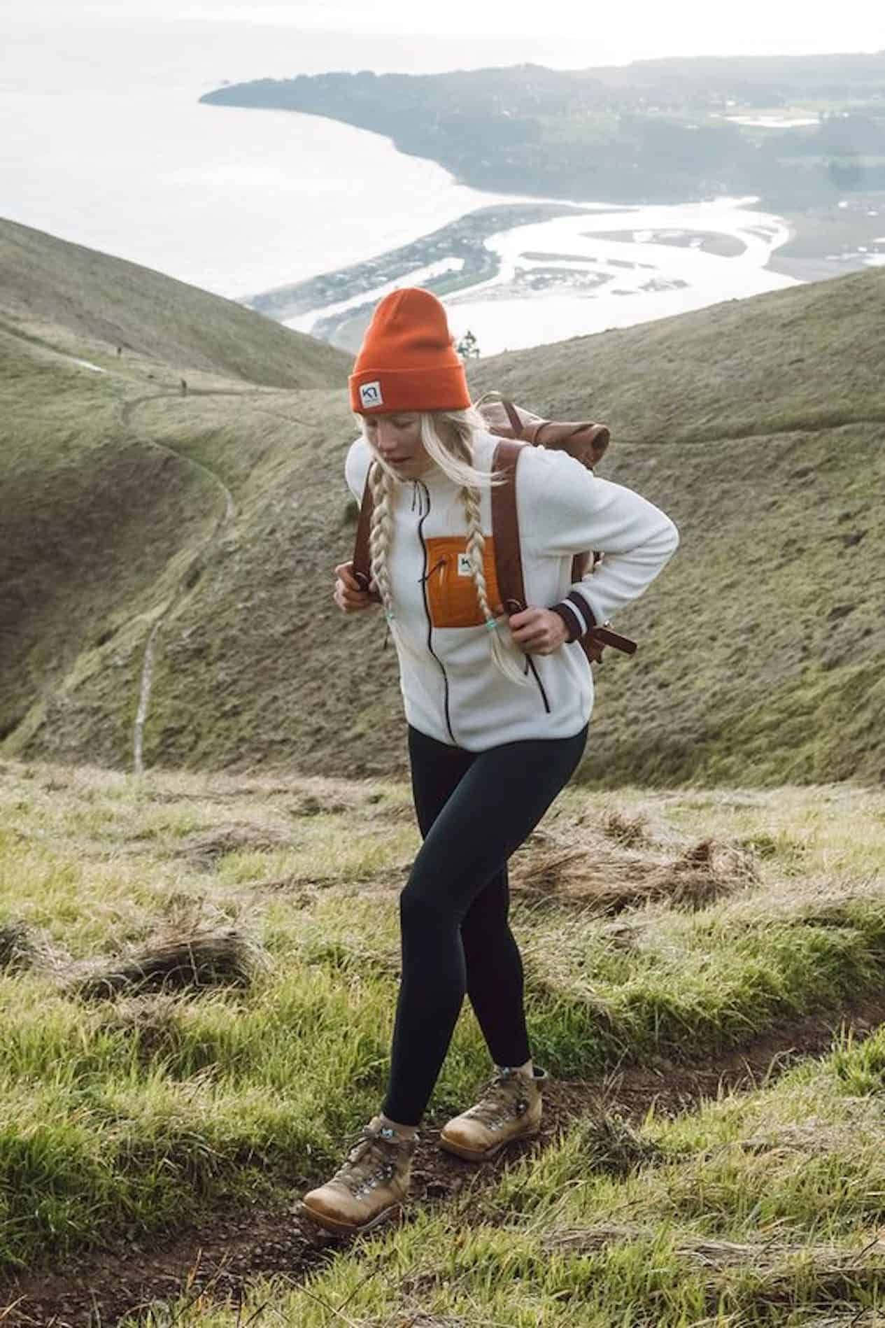 Winter Hiking 桌布 壁紙 — 一名戴著橙色冷帽、身穿白色連帽衛衣的女子，在俯瞰蜿蜒海岸灣的草地上行山。