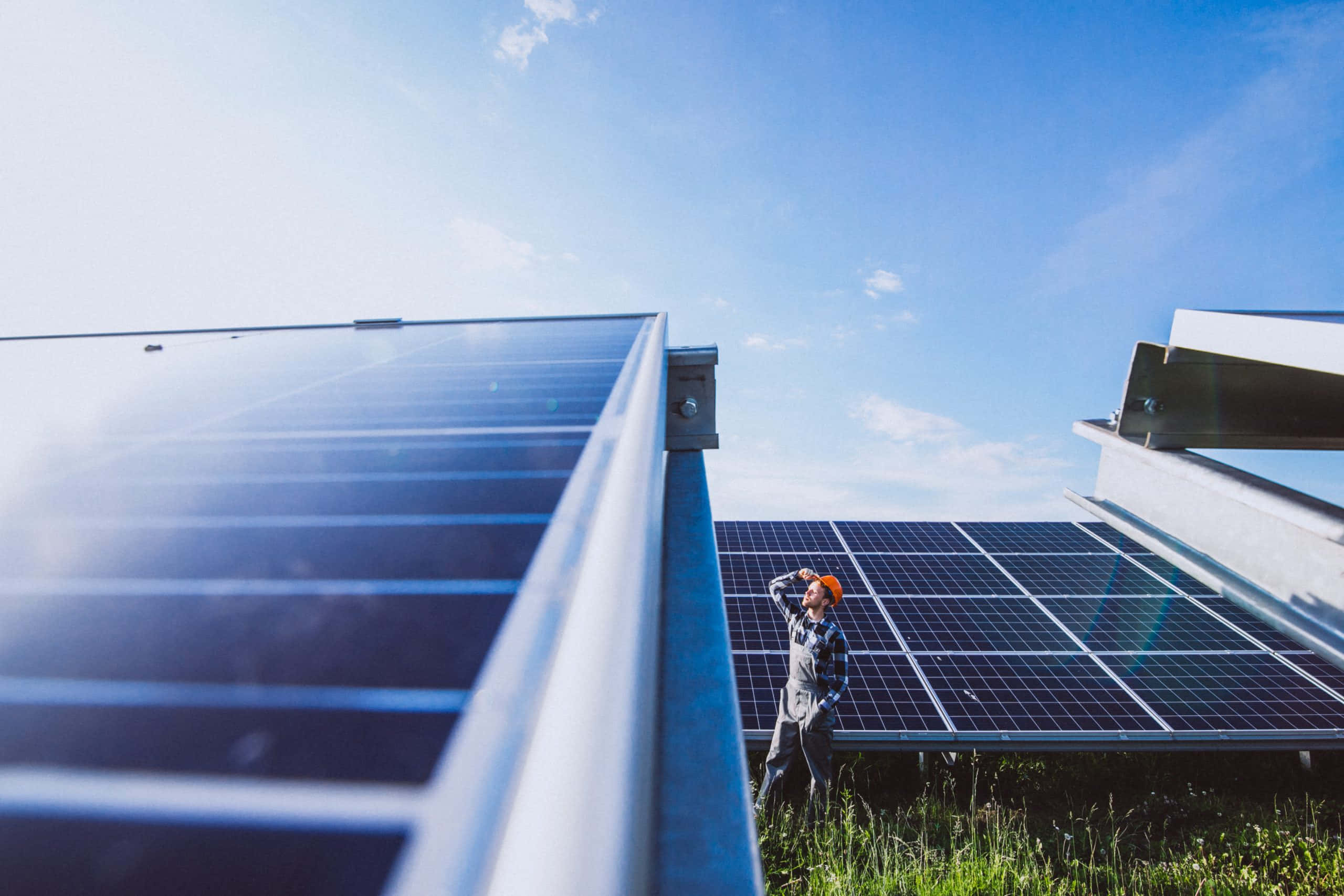 Solar Panel 桌布 壁紙 — 一名戴著橙色安全帽的技術員站在廣闊的太陽能板下，天空湛藍，白雲點點。