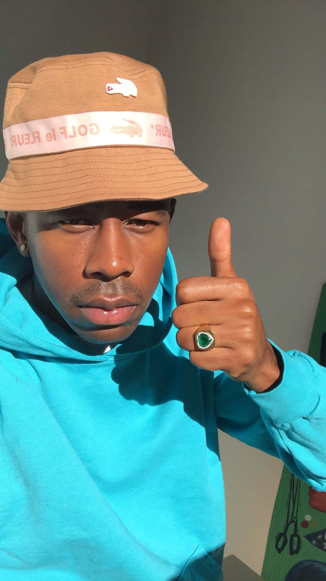 tyler the creator 桌布 壁紙 — 一名戴著米色漁夫帽和亮藍色連帽衫的男子，戴著一個大綠戒指，豎起大拇指。