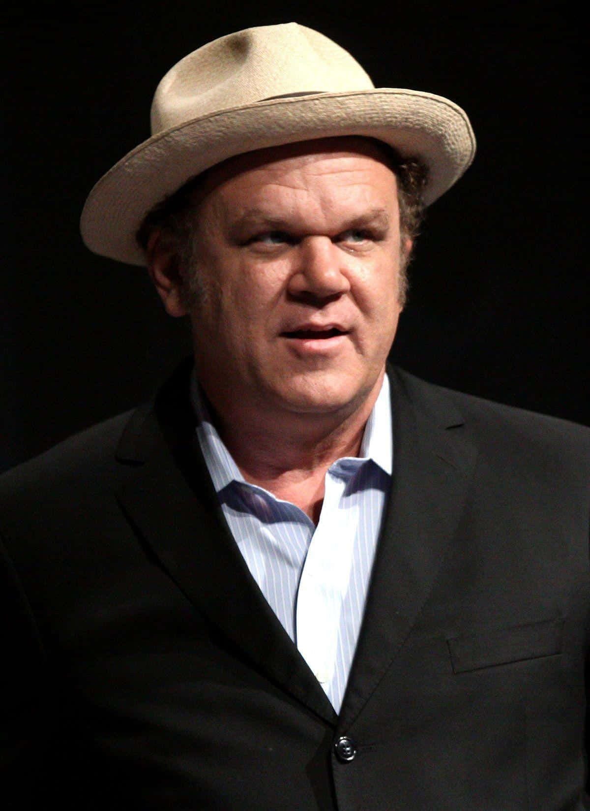 John C. Reilly 桌布 壁紙 — 一名戴著米色軟呢帽、身穿黑色西裝的男子，在聚焦燈光下站在深色背景前。