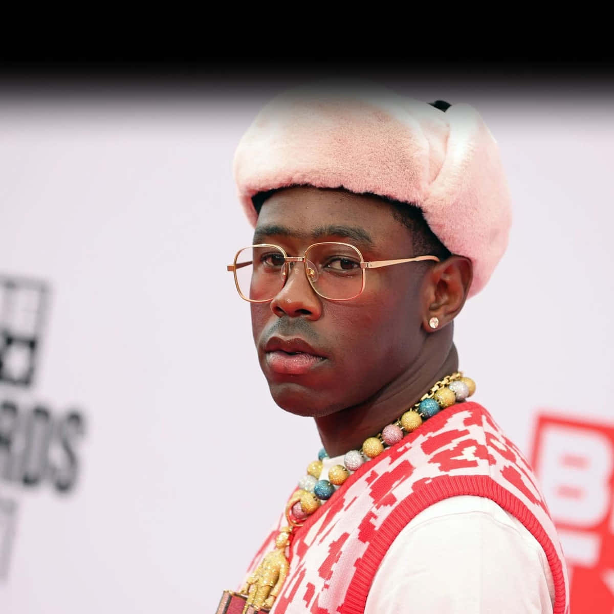 tyler the creator 桌布 壁紙 — 一名戴著粉紅色絨毛帽和金邊眼鏡的男子，身穿彩色珠鏈和圖案背心，在紅地毯上擺姿勢。