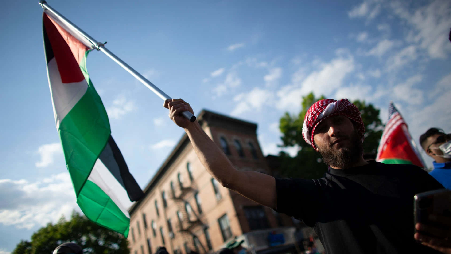 Palestine 桌布 壁紙 — 一名戴著紅白頭巾的男子在晴朗藍天白雲下高舉巴勒斯坦旗幟。