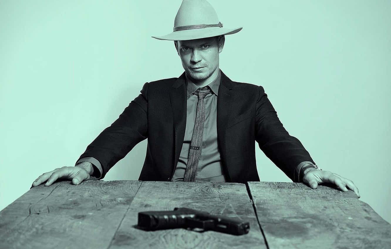 timothy olyphant 桌布 壁紙 — 一名戴著紳士帽、身穿西裝的男子，在淺綠色背景前，俯身在一張木桌上，桌上放著一把手槍。