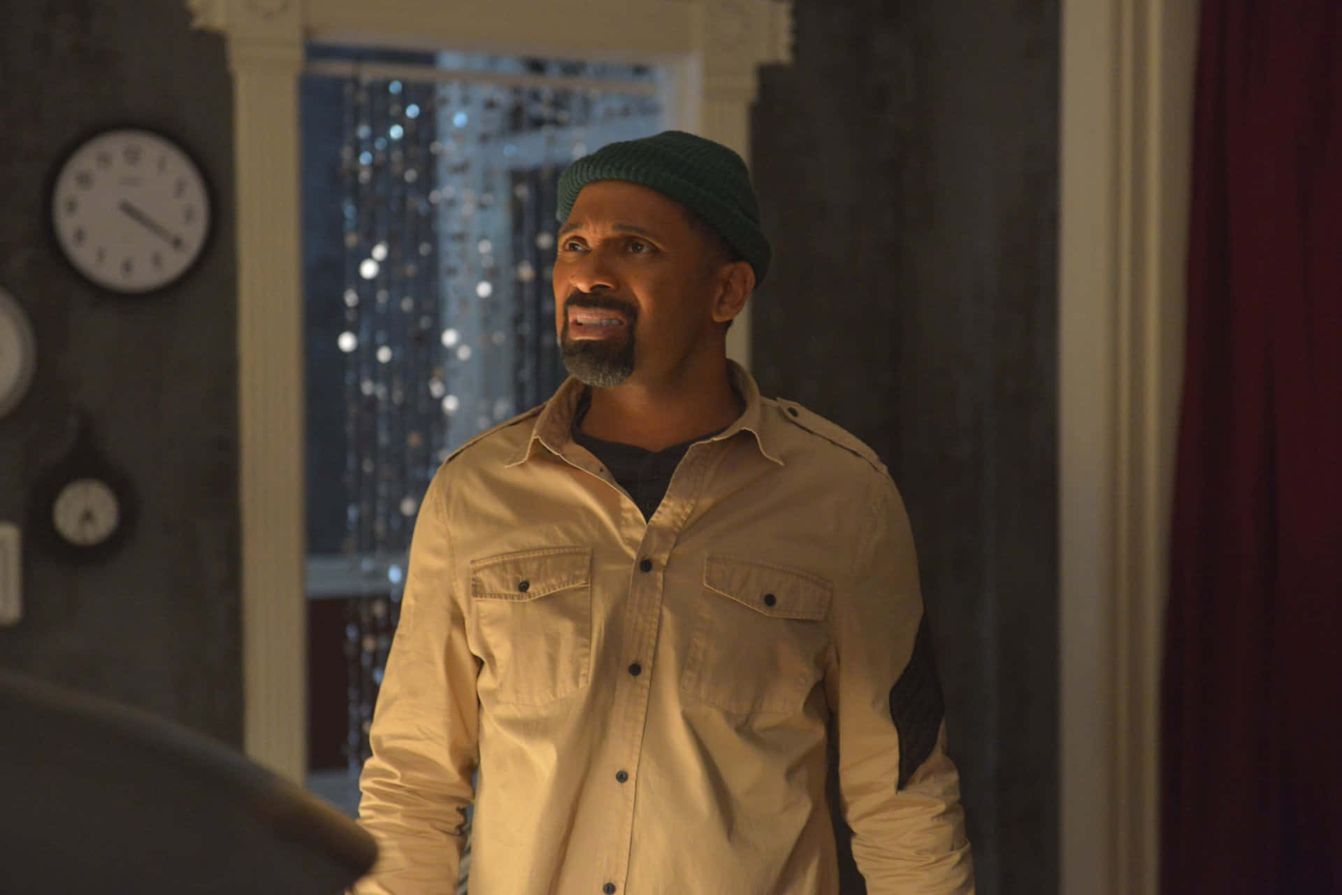 Mike Epps 桌布 壁紙 — 一名戴著綠色冷帽、身穿米色襯衫的男子站在光線昏暗的房間裡，身後有時鐘和窗簾。