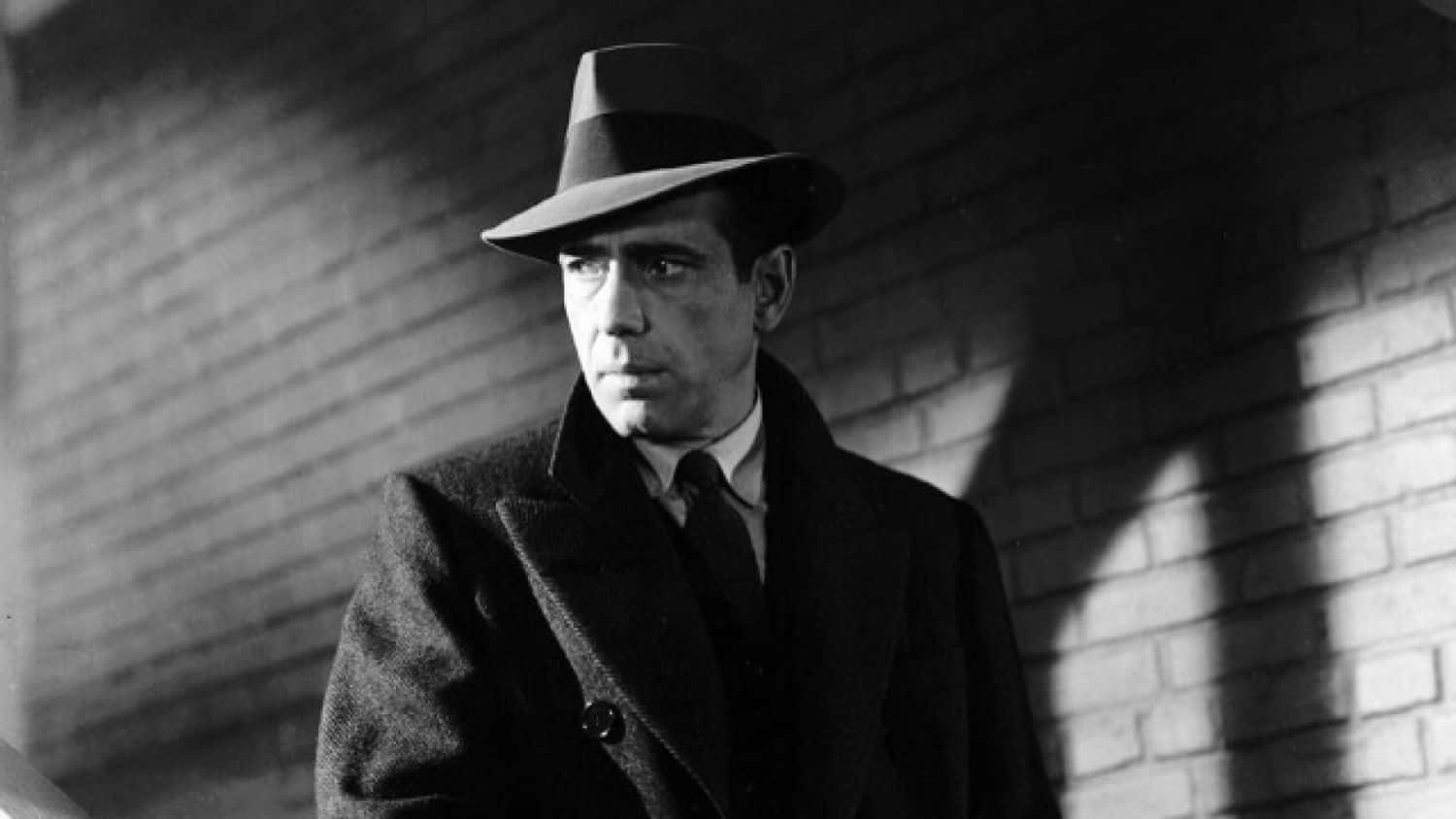Film Noir 桌布 壁紙 — 一名戴著費多拉帽和穿著風衣的男子，在戲劇性的明暗對比燈光下靠著磚牆。