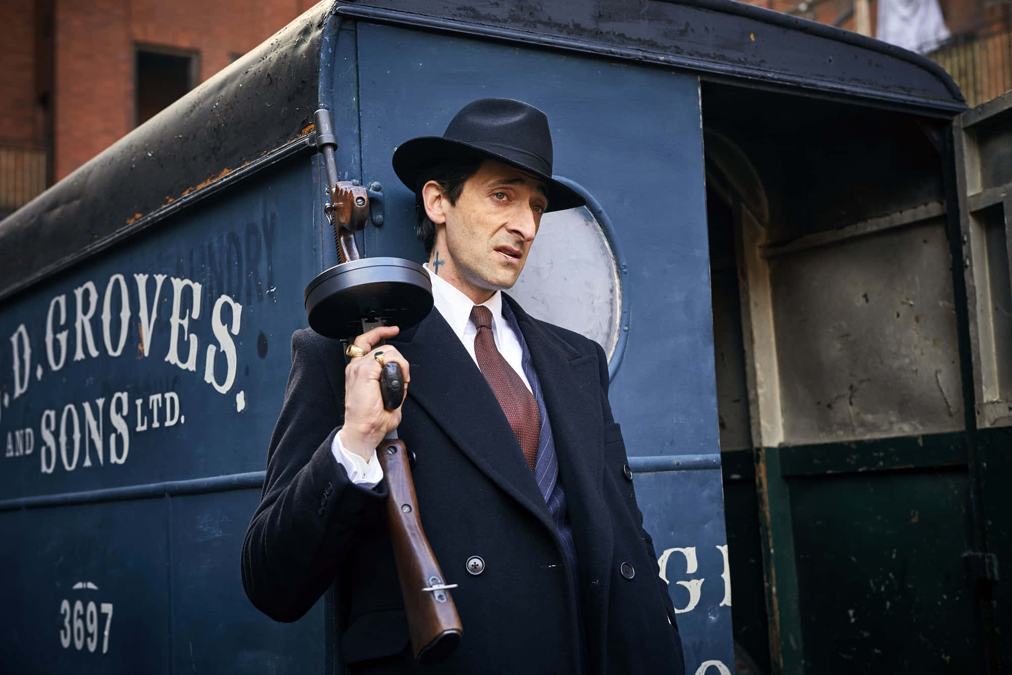 Peaky Blinders 桌布 壁紙 — 一名戴著軟呢帽、身穿西裝的男子，在城市環境中拿著一把復古霰彈槍，旁邊是一輛藍色送貨車。