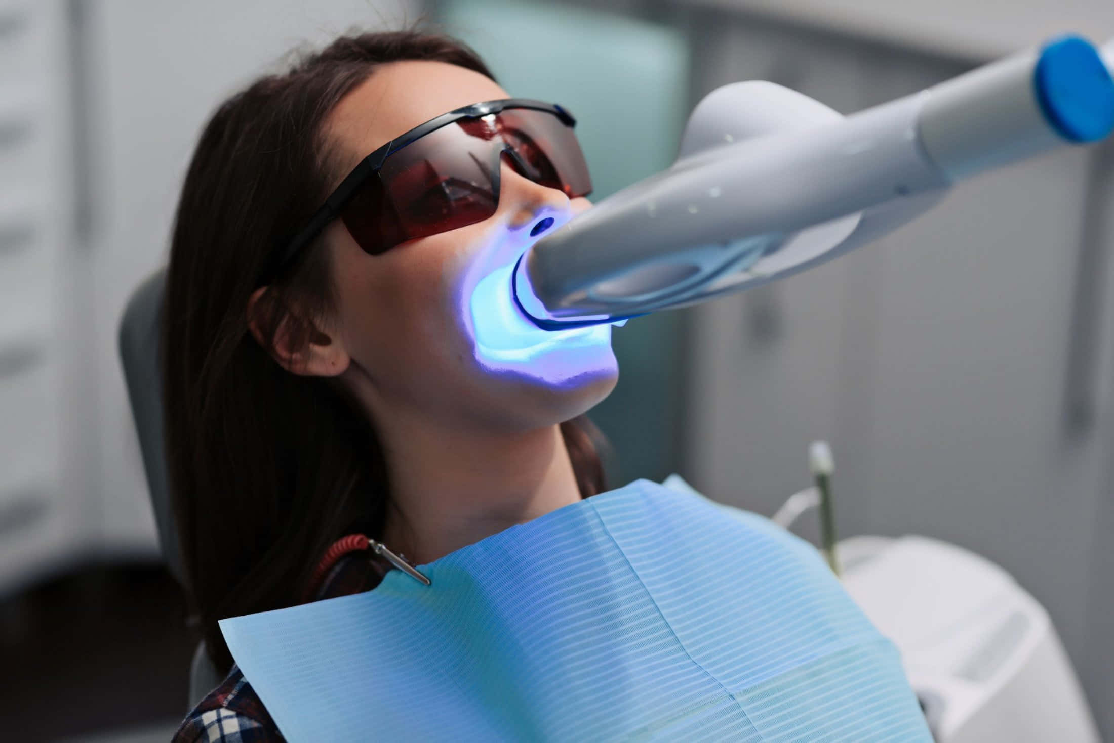 Teeth Whitening 桌布 壁紙 — 一名戴著防護眼鏡的女性在臨床環境中接受明亮的藍光牙科治療。
