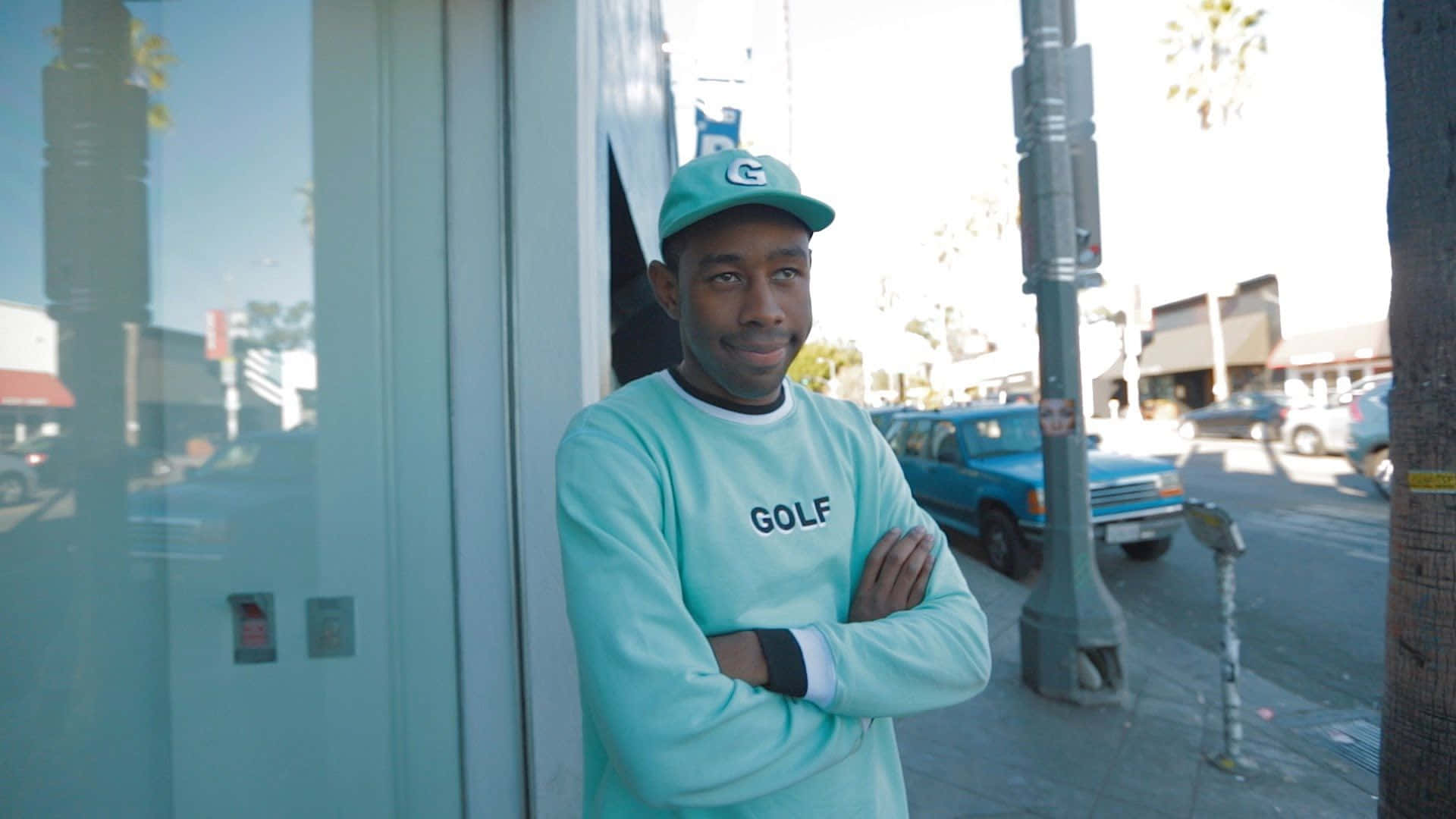 tyler the creator 桌布 壁紙 — 一名戴著青綠色帽子、身穿相配的「GOLF」運動衫的男子，在陽光明媚的城市人行道上自信地交叉雙臂站立。