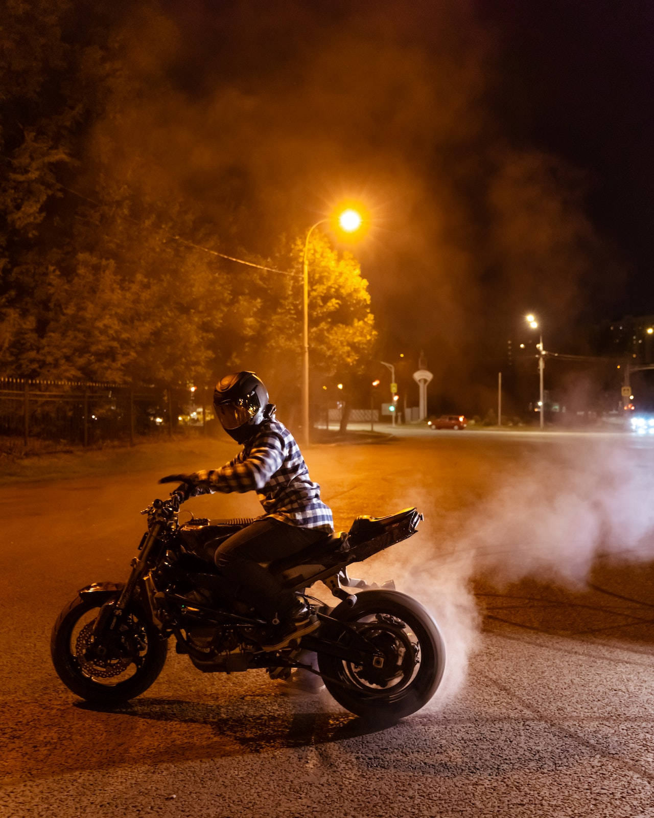 Bike Rider 桌布 壁紙 — 一名戴著頭盔、身穿格仔恤衫的電單車手在夜晚燒胎，在發光的街燈下揚起煙霧。