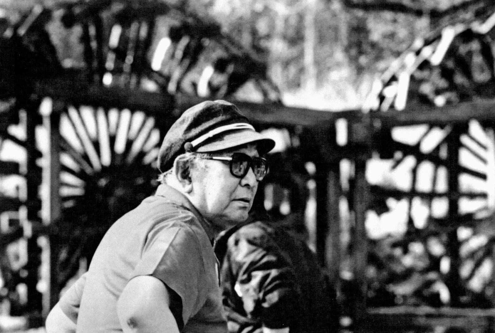 Akira Kurosawa 桌布 壁紙 — 一名戴著鴨舌帽和太陽眼鏡的男子，在一個大型木製水車旁若有所思地凝視著黑白畫面。