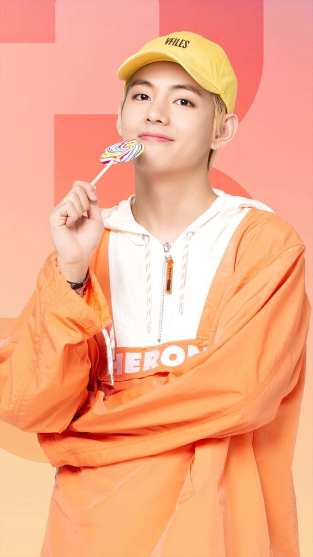 Taehyung Cute 桌布 壁紙 — 一名戴著黃色鴨舌帽、身穿橙色連帽衛衣的年輕男子，在柔和的粉紅色幾何背景前拿著一支彩色棒棒糖。