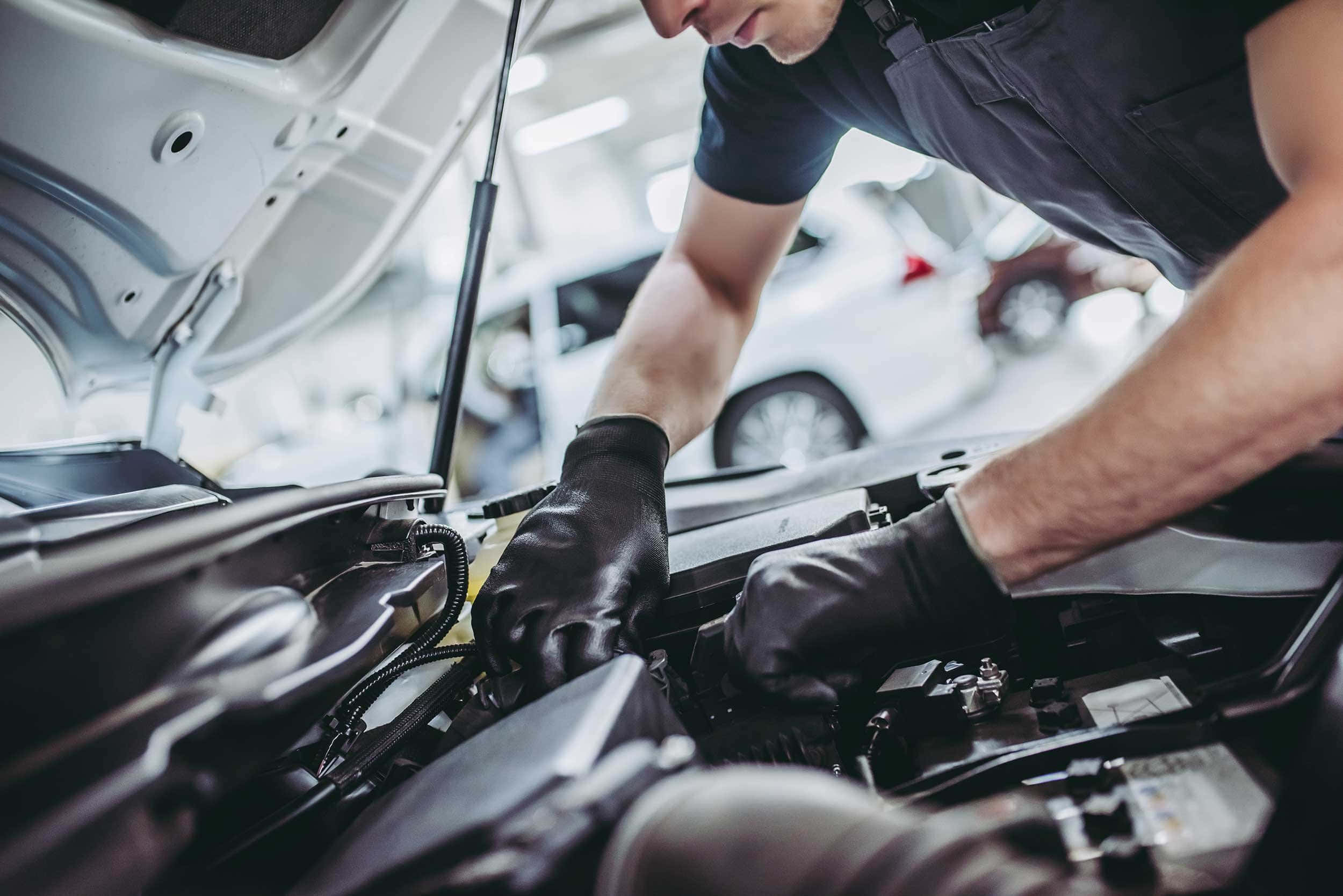 Car Repair 桌布 壁紙 — 一名戴著黑色手套的技師在明亮的汽車維修廠中，仔細地在汽車引擎蓋下工作。
