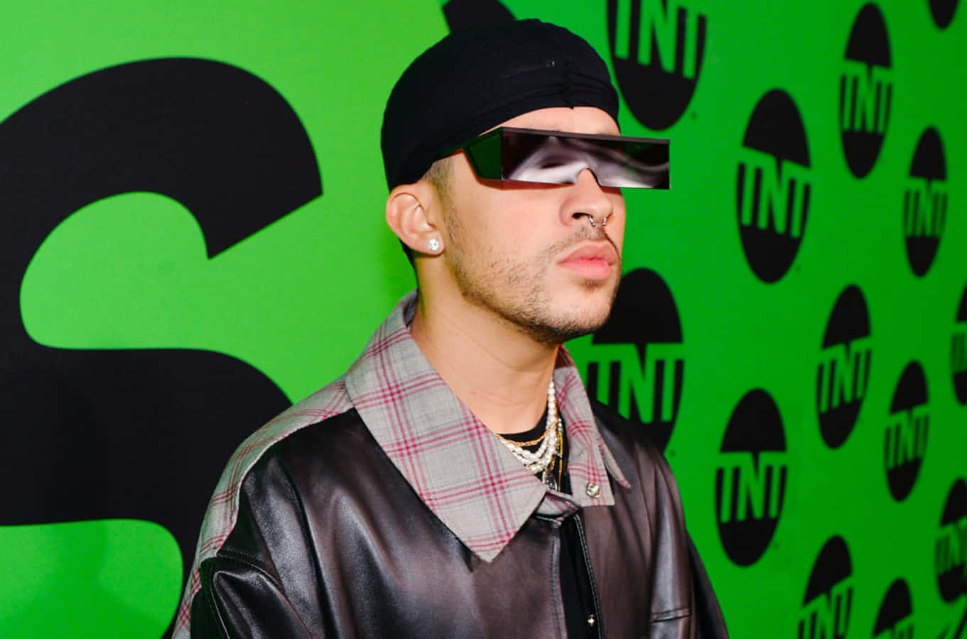 Bad Bunny 桌布 壁紙 — 一名戴著黑色鴨舌帽和未來感太陽眼鏡的男子，在帶有重複黑色標誌的鮮綠色牆前擺姿勢。