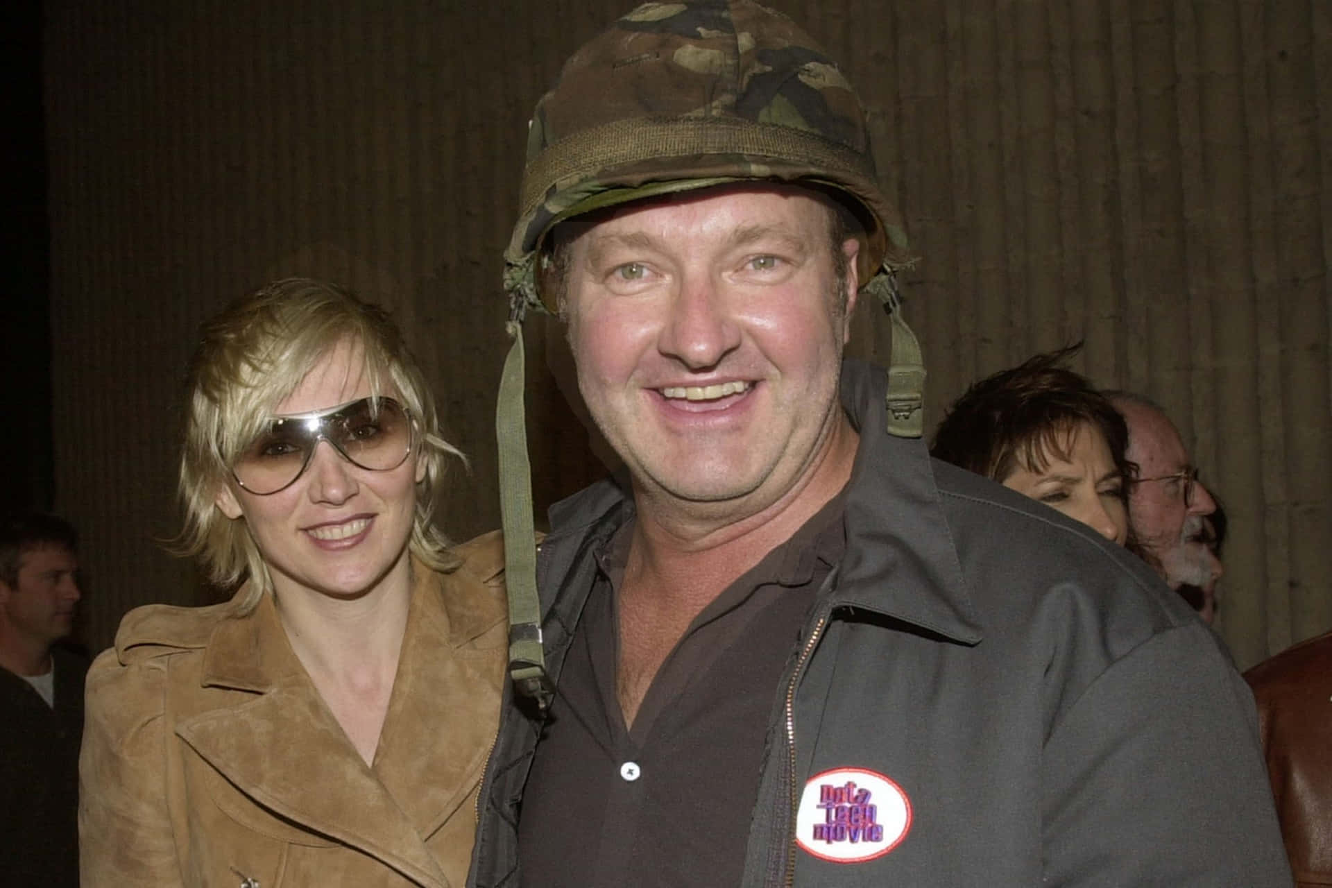 Randy Quaid 桌布 壁紙 — 一名戴迷彩頭盔的男子和一名戴太陽眼鏡的女子在室內活動中微笑，背景是帶紋理的牆壁。