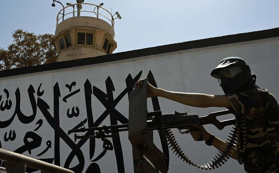 Taliban 桌布 壁紙 — 一名戴面具的武裝警衛身穿迷彩服，在晴朗天空下從帶有阿拉伯書法的牆後瞄準機槍。